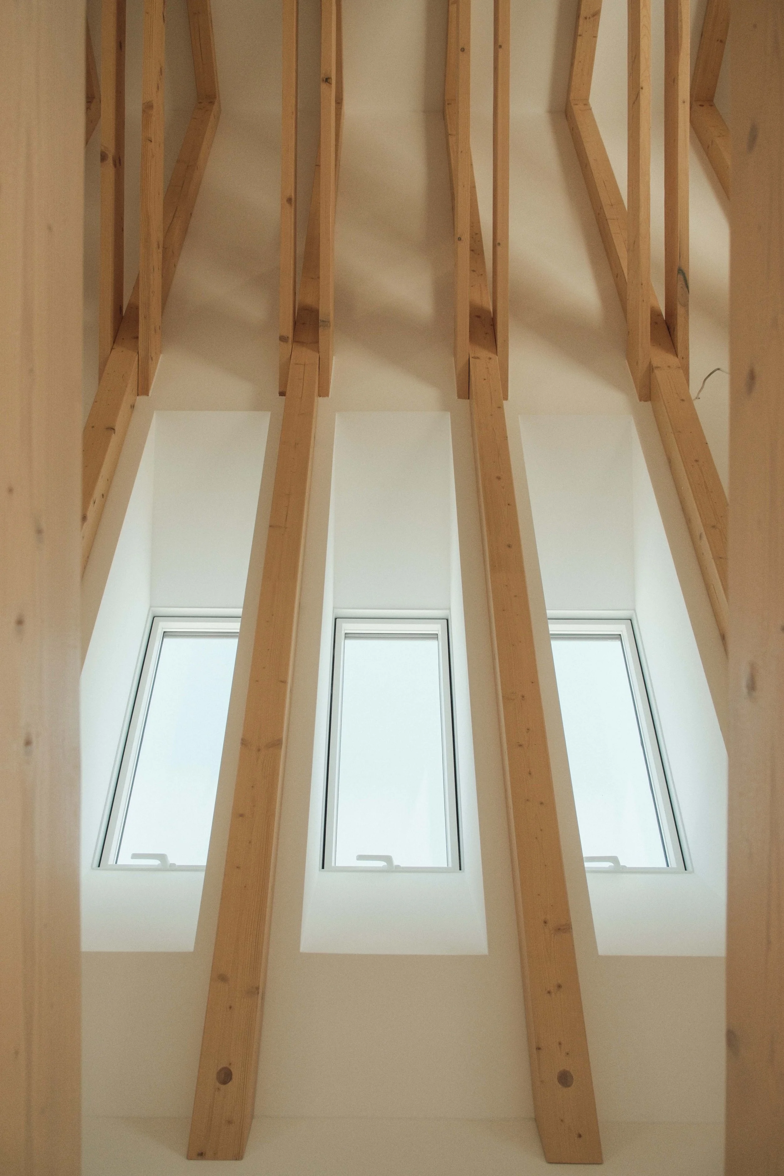 The image shows a view of an attic with unfinished wooden beams and two vertical windows letting in natural light from outside.