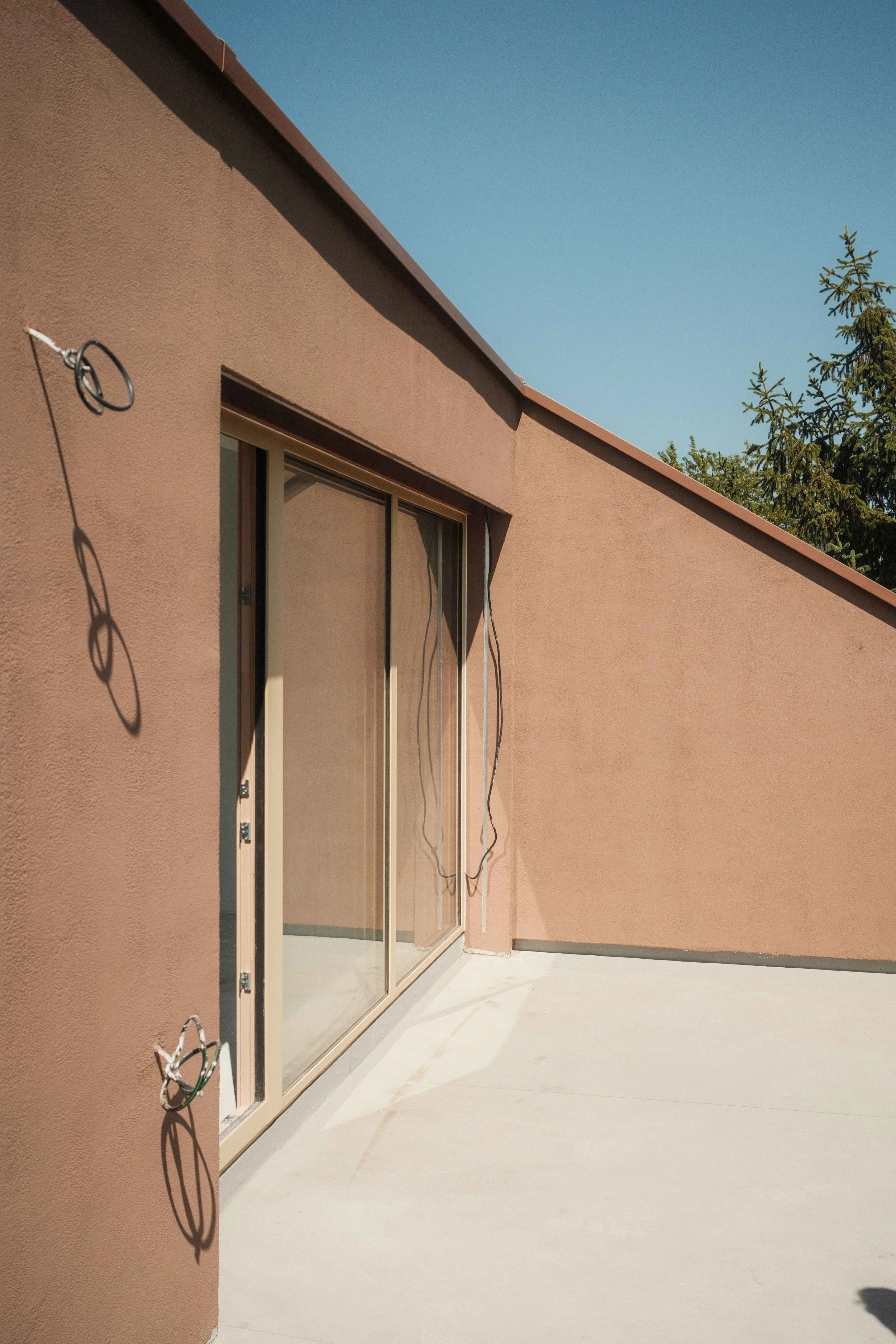 A pinkish-beige exterior wall with a sliding glass door and some wires hanging loosely on the wall, under a clear blue sky. Part of a green tree is visible in the background.