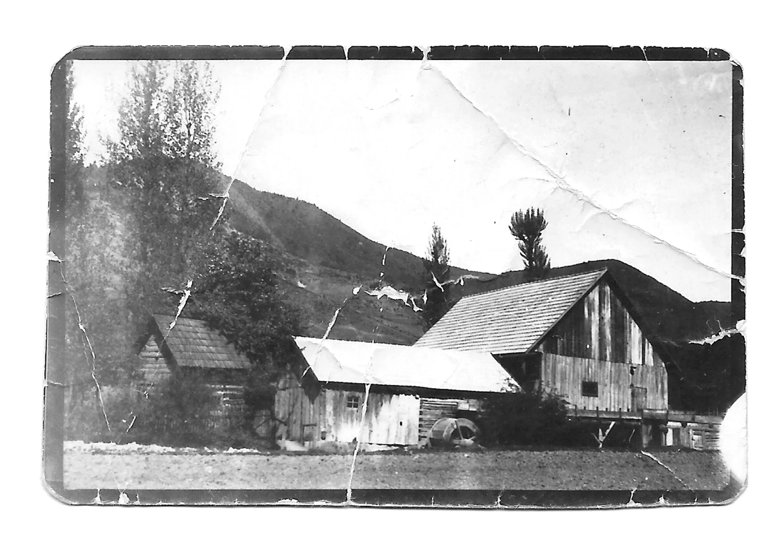 Black-and-white photograph of a rural scene with hillside, trees, and a barn-like building with a slanted roof, seen through a cracked, weathered window.