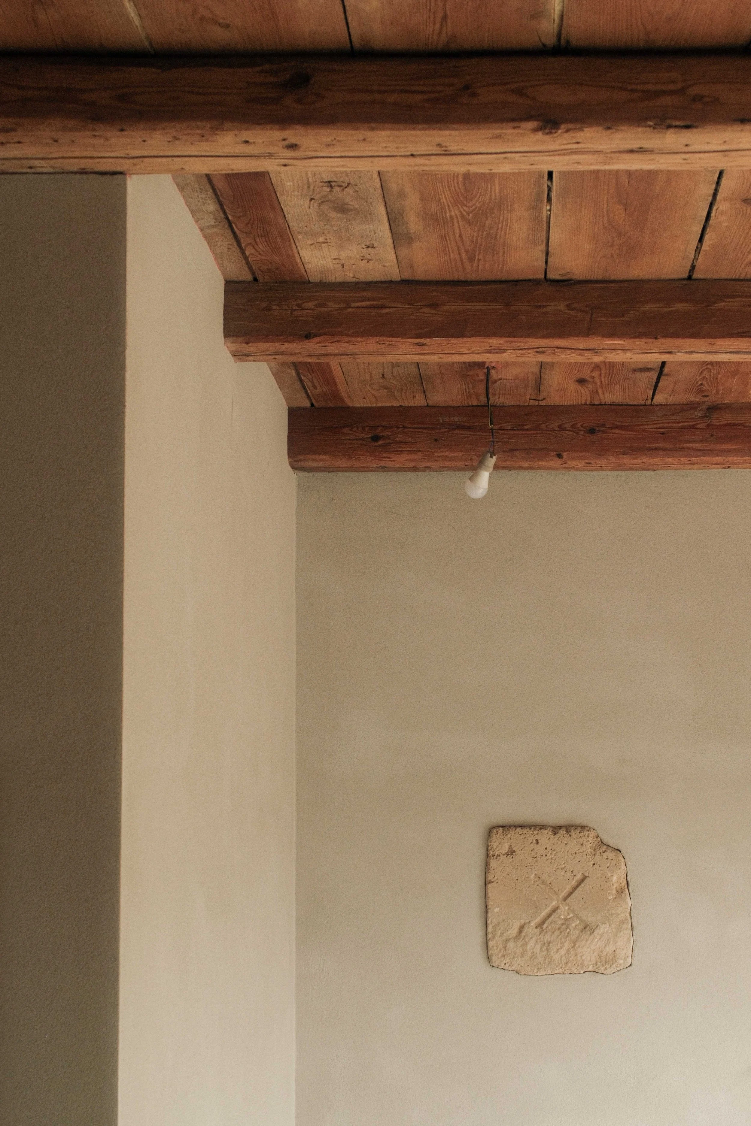 A ceiling with wooden beams, a hanging light bulb, and a portion of a brick or stone wall.