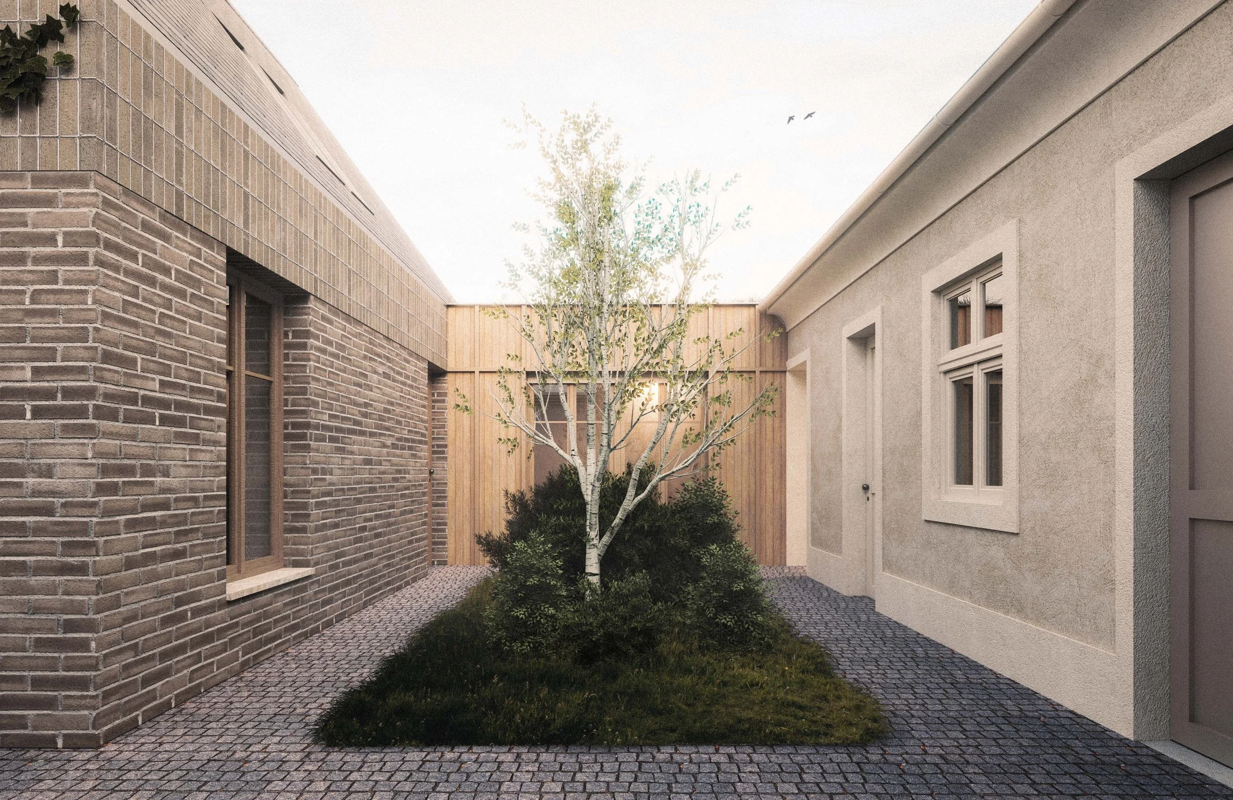 A small courtyard with a young tree surrounded by shrubbery, enclosed by brick and stucco buildings, and paved with gray cobblestone.
