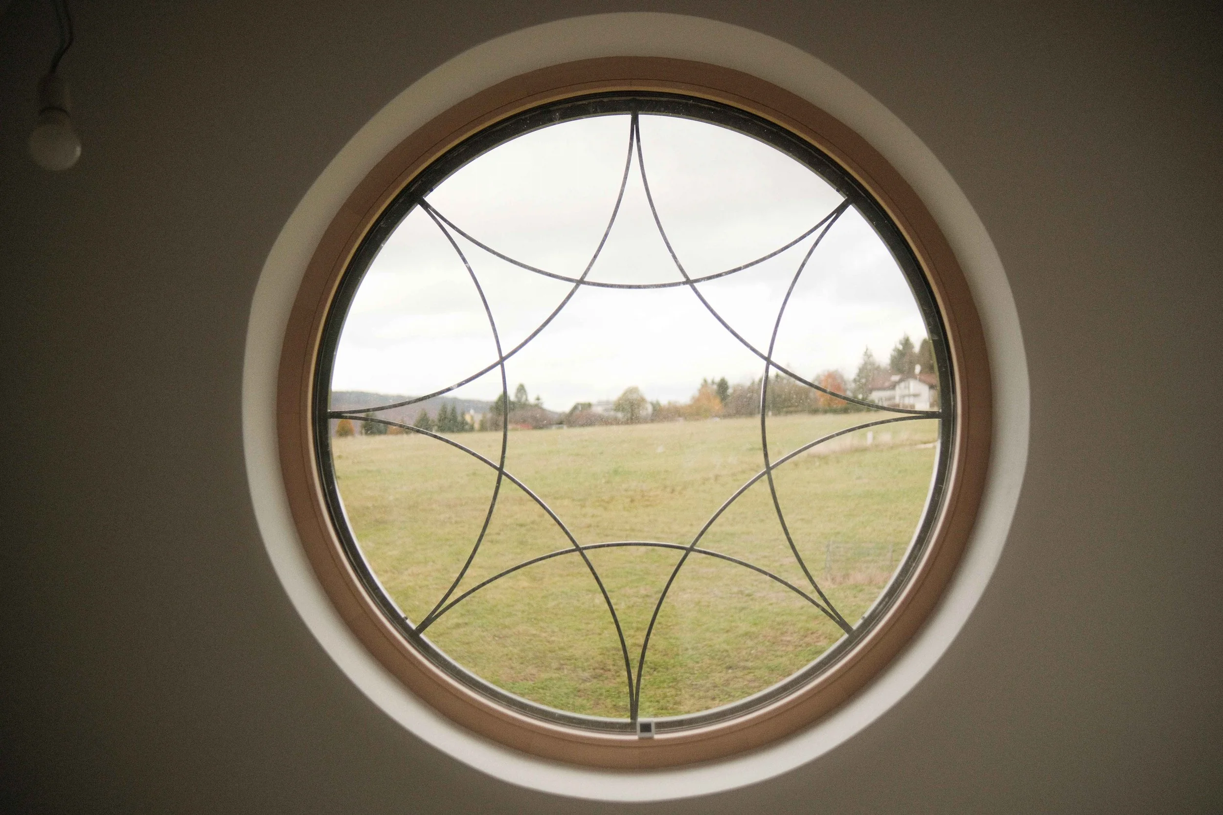A round window with decorative black metal grille design, showing an outdoor landscape with grass, trees, and houses under a cloudy sky.
