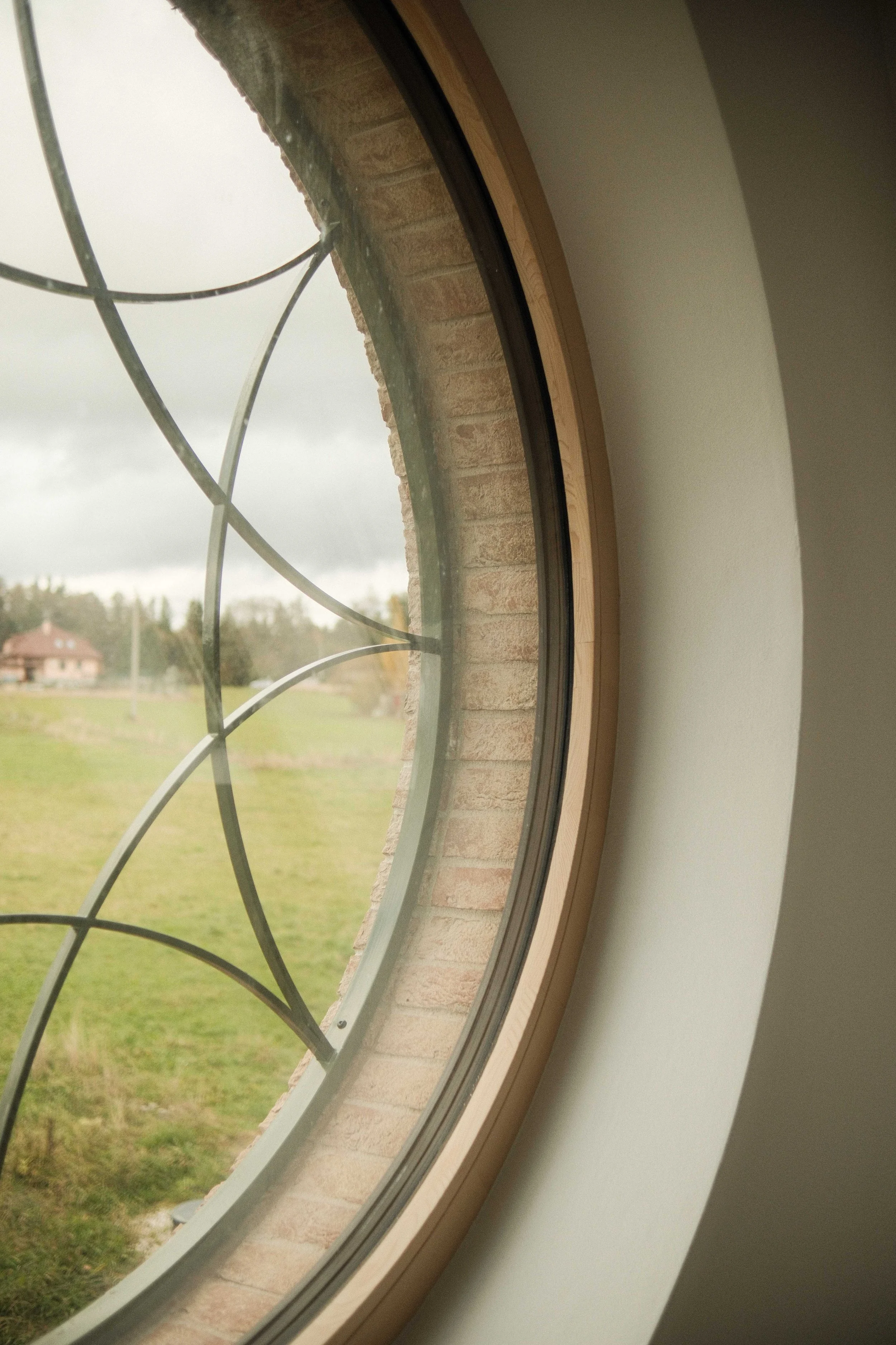A view through a circular window with a metal grid into a green outdoor landscape with a house in the background, cloudy sky, and trees.