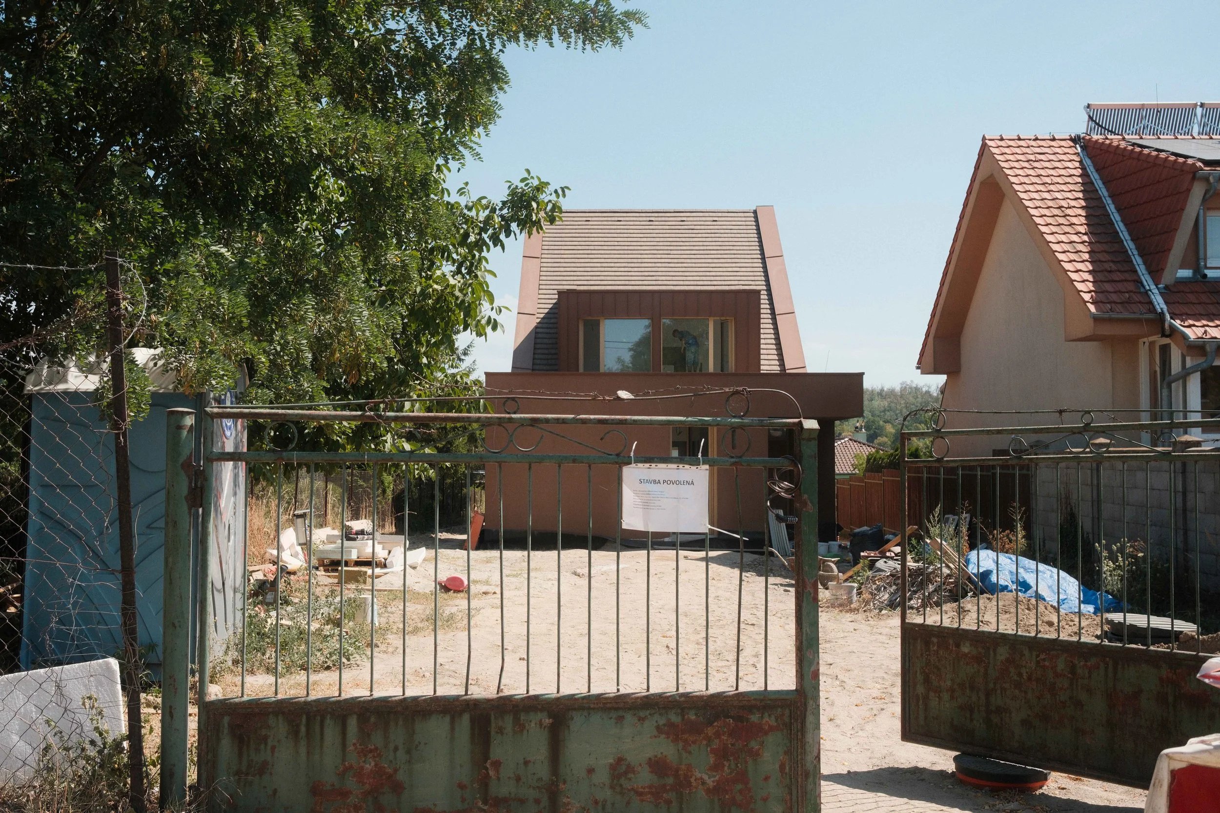 View of a construction site behind a rusty gate, with a newly built modern house and another house with a red-tiled roof in the background. Equipment and materials are scattered on the ground.