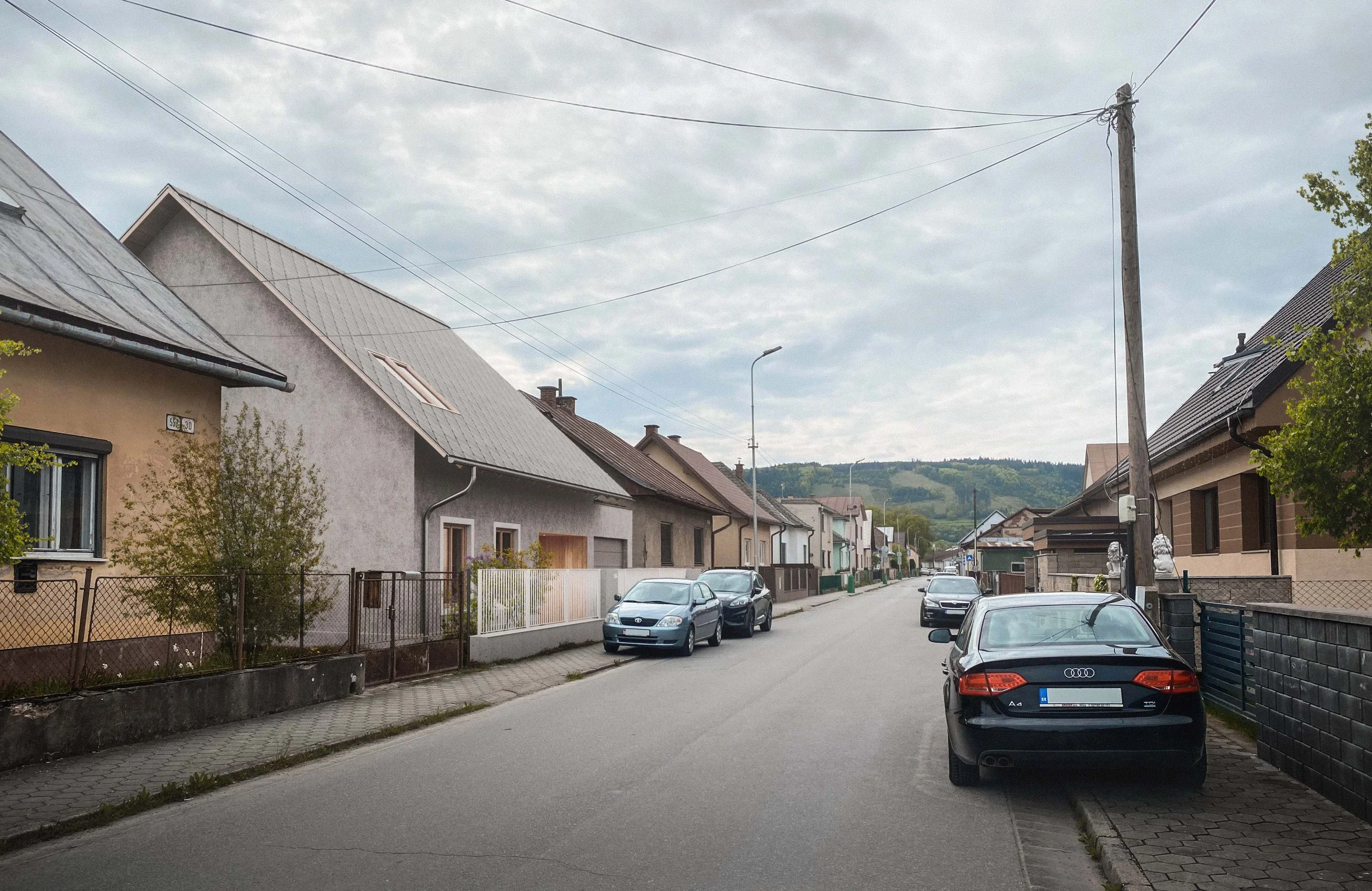 A quiet residential street with houses on both sides, parked cars, utility poles, and a cloudy sky above.