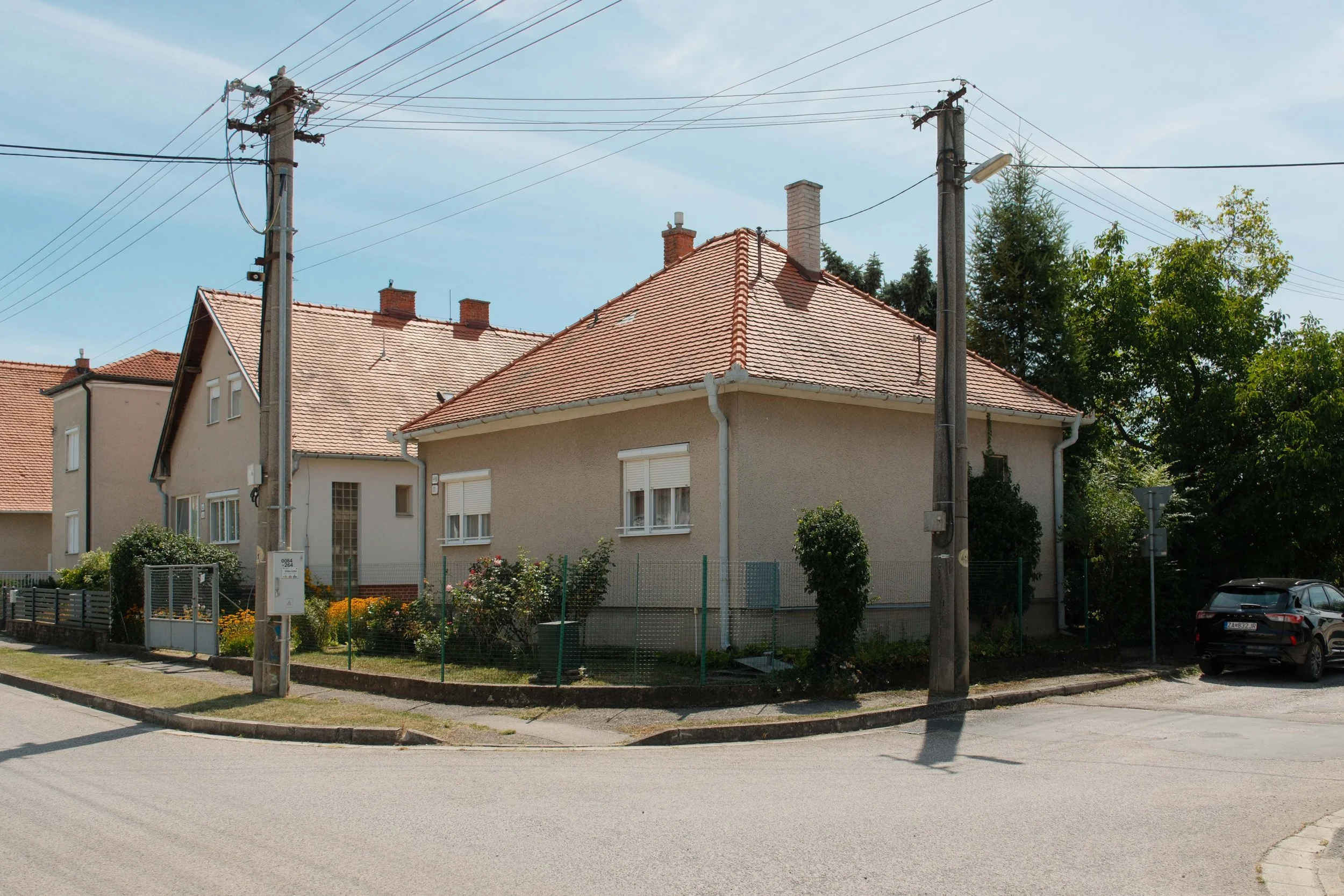 A residential neighborhood scene with two houses featuring red-tiled roofs, surrounded by a small garden enclosed by a green fence. Two utility poles with multiple wires are in the foreground, and a street with a parked car is visible. Trees and a clear blue sky complete the background.