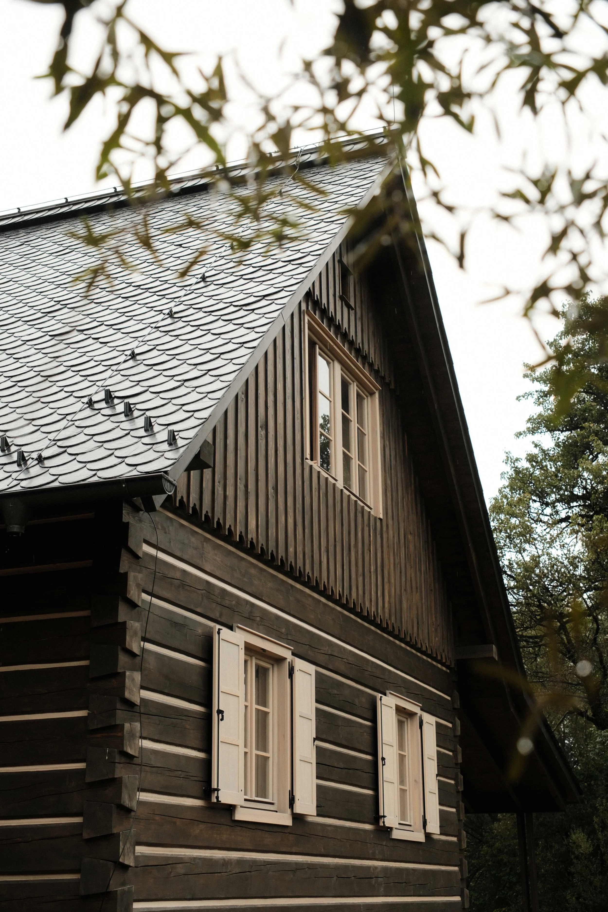 A multi-story wooden house with a sloped roof covered in grey shingles, surrounded by trees, with white window shutters and a small balcony.