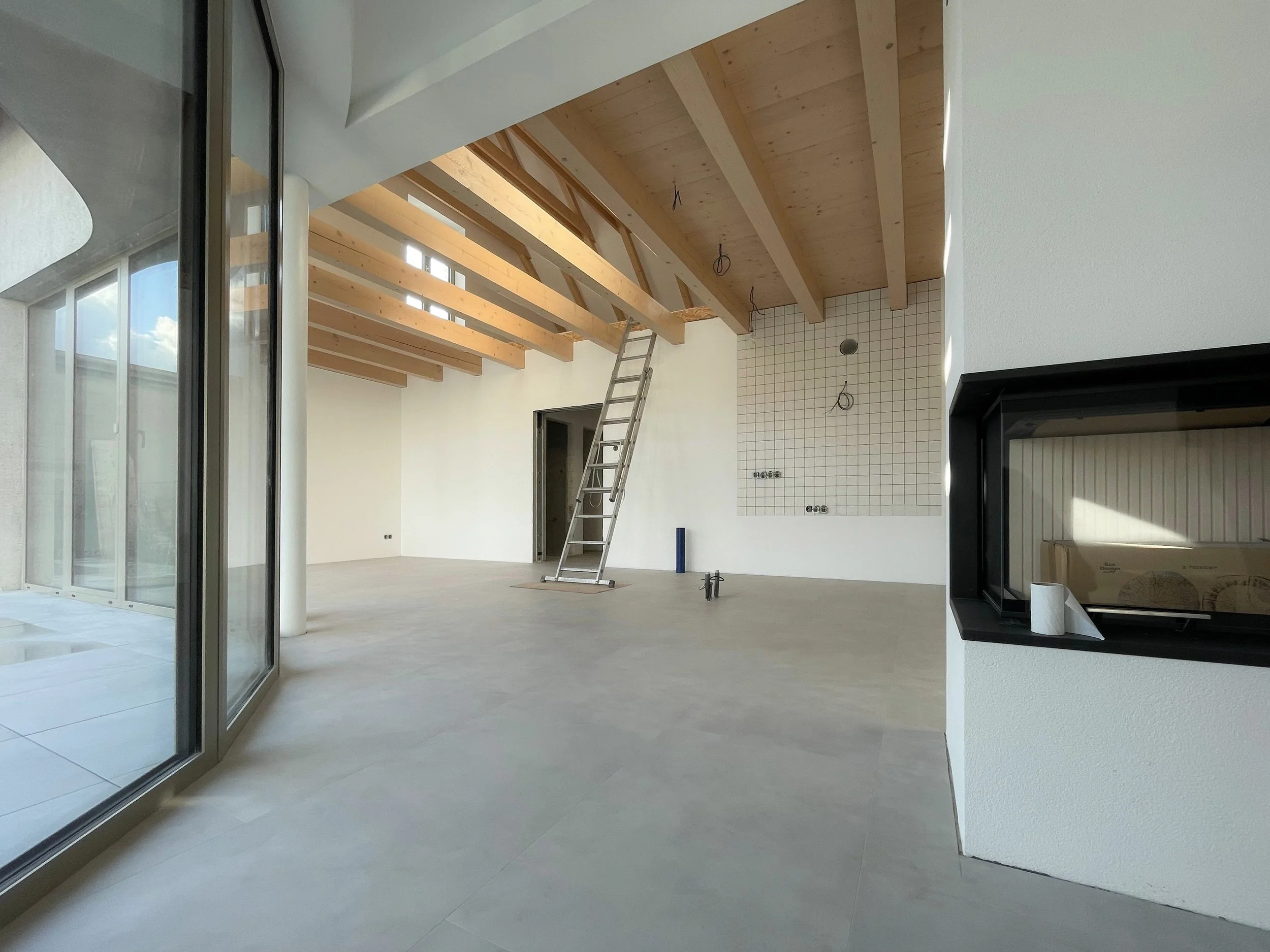 Modern living space under construction with exposed wooden beams, a ladder, and a tiled wall area.