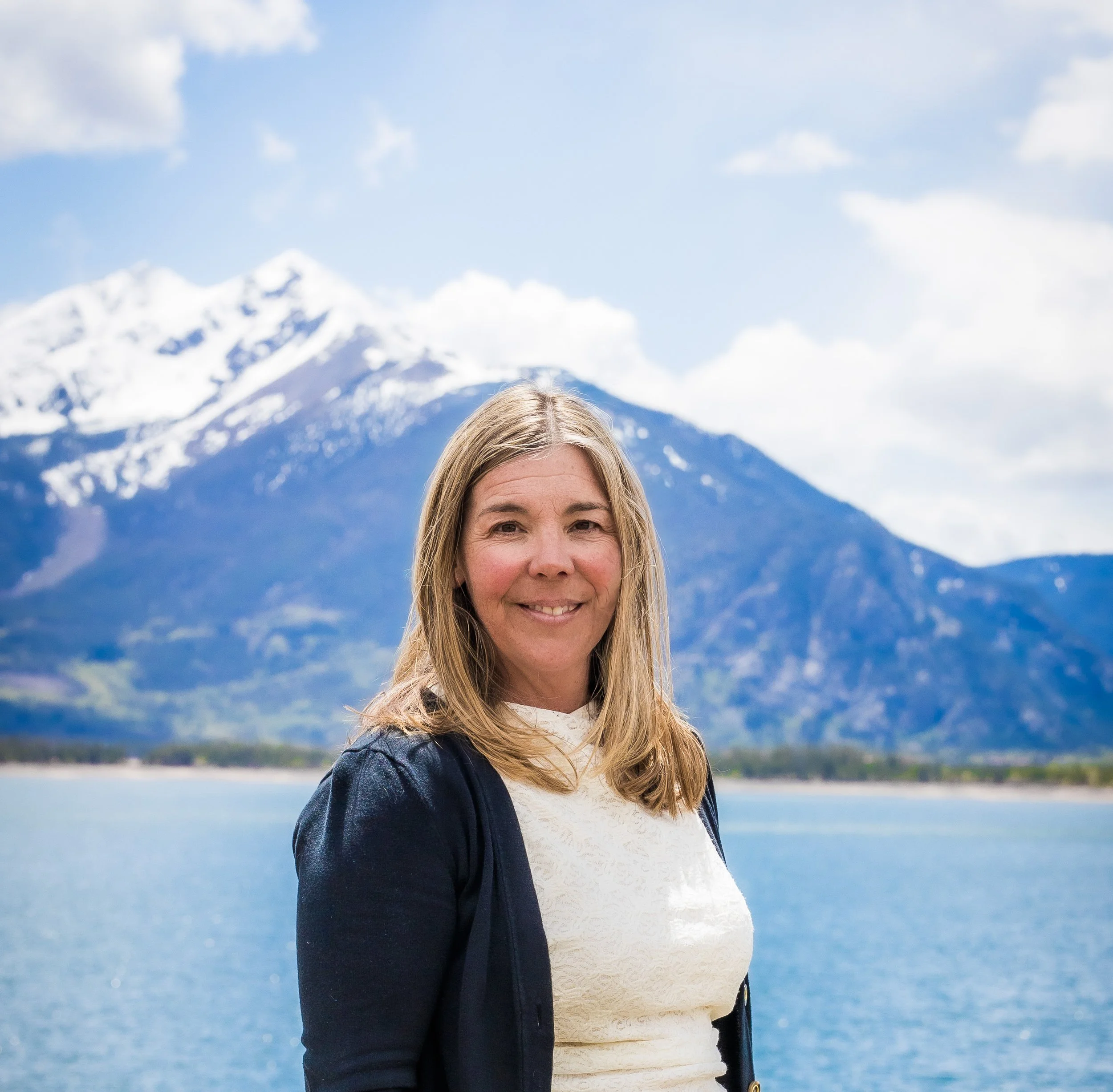 A woman with blonde hair smiling in front of snowy mountains and a lake with blue sky and clouds.