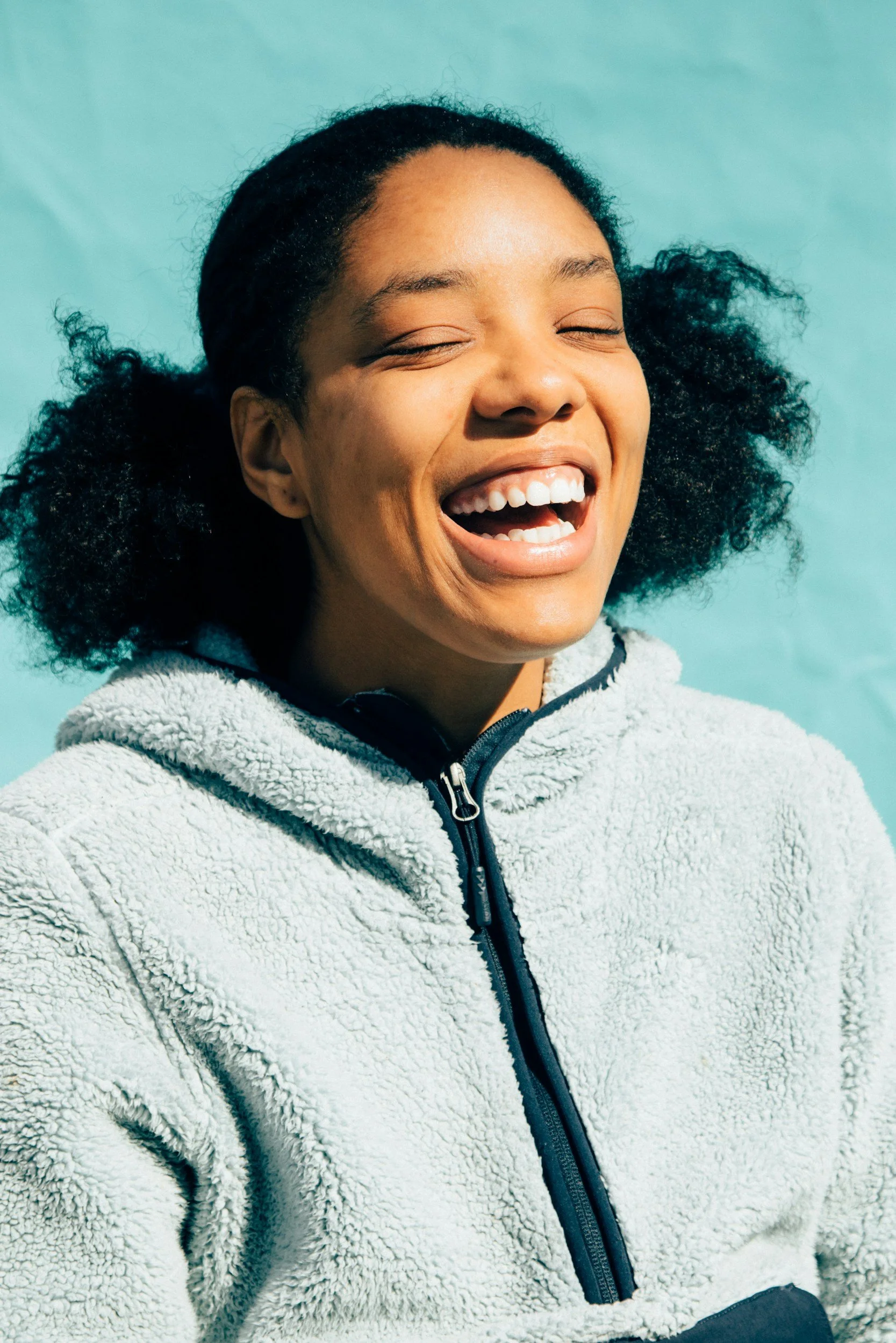 Close-up of a woman with dark, curly hair smiling and laughing, wearing a light gray fleece jacket against a teal background.