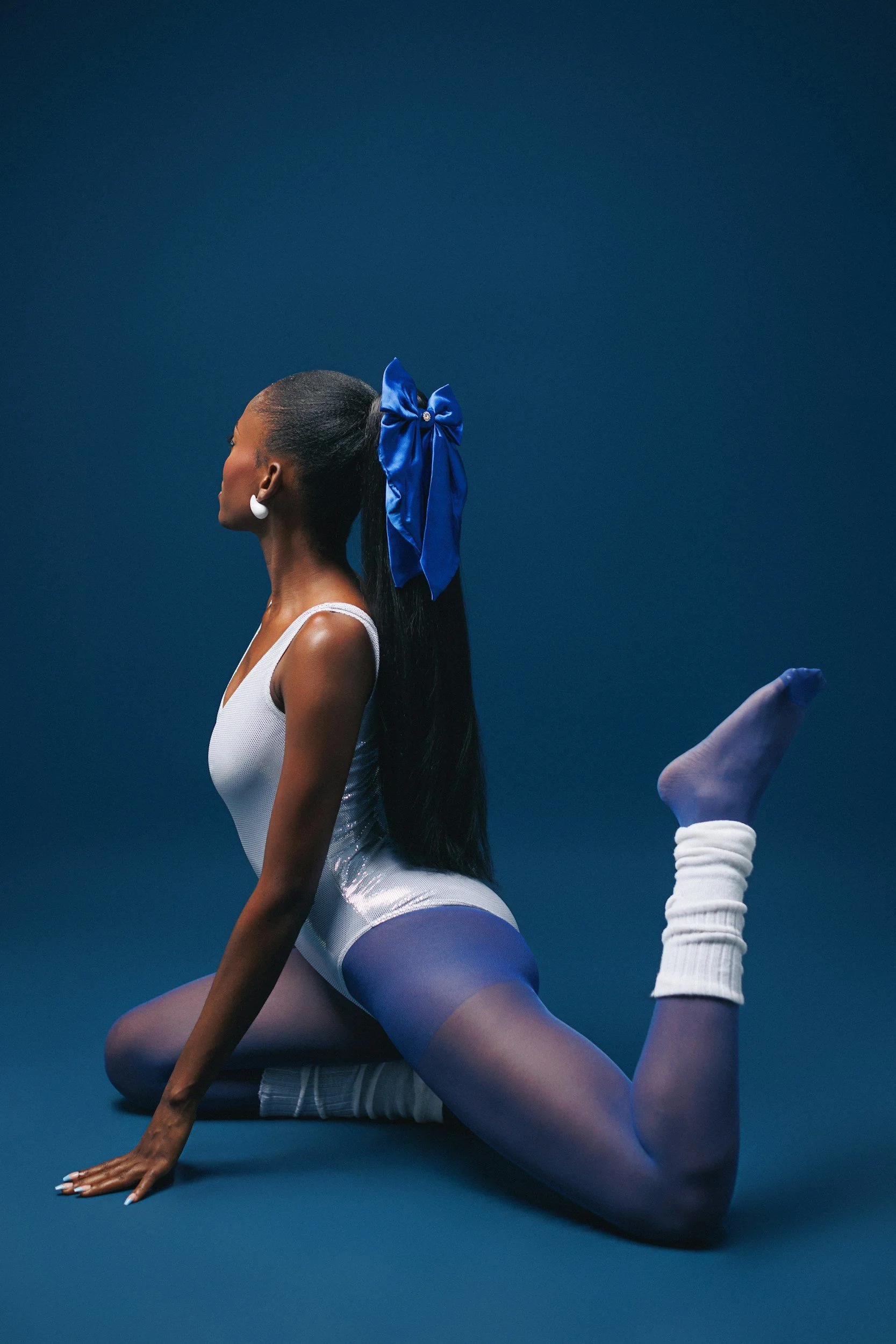 A woman in a white leotard with a blue satin bow in her hair and white earrings poses in a yoga stretch on a blue background. She is practicing a yoga pose with one knee on the ground, one arm extended on the ground, and the other leg bent upward.