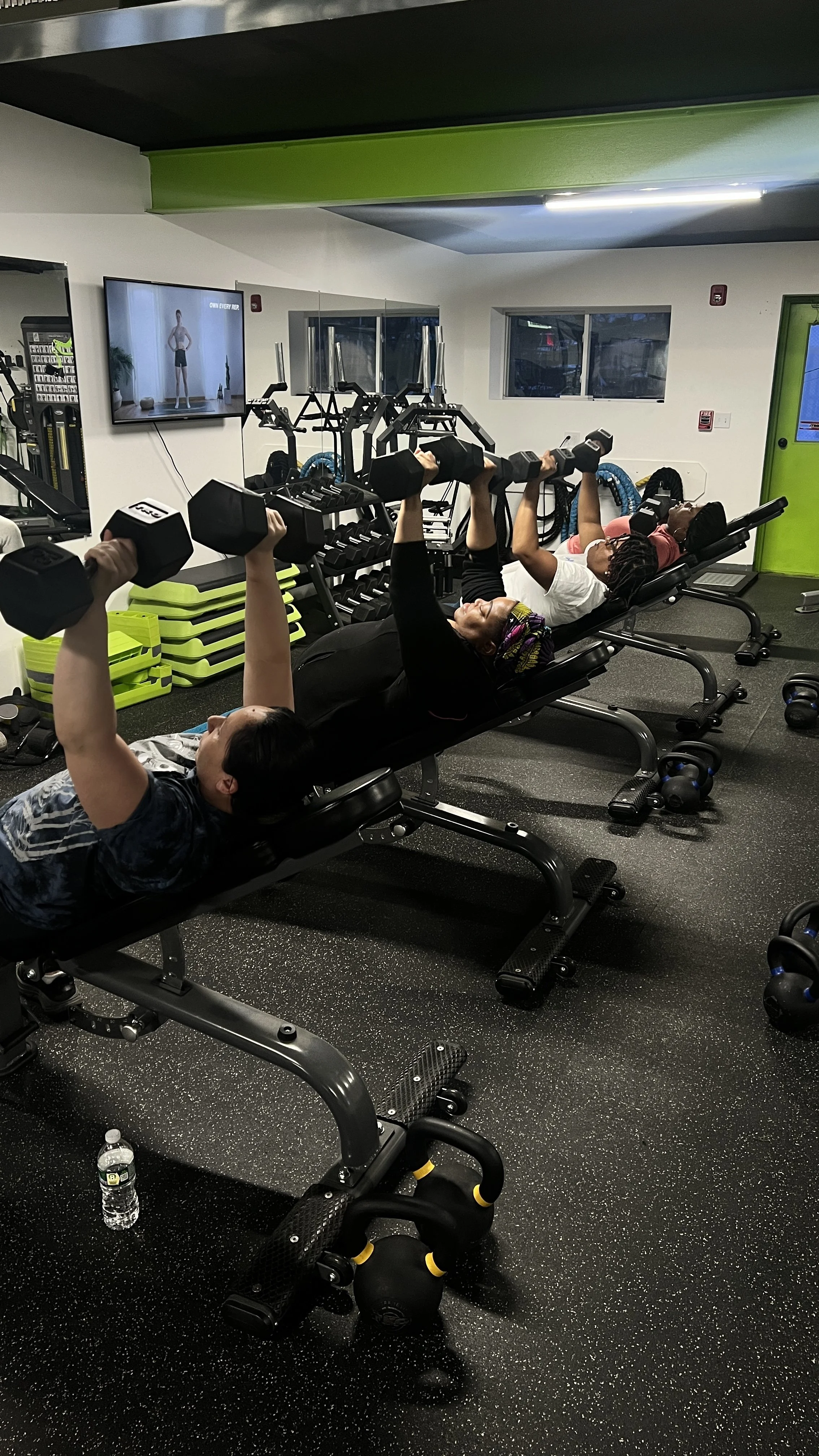 People lying on workout benches lifting dumbbells in a gym, with a TV screen showing a workout instructor and green storage bins in the background.