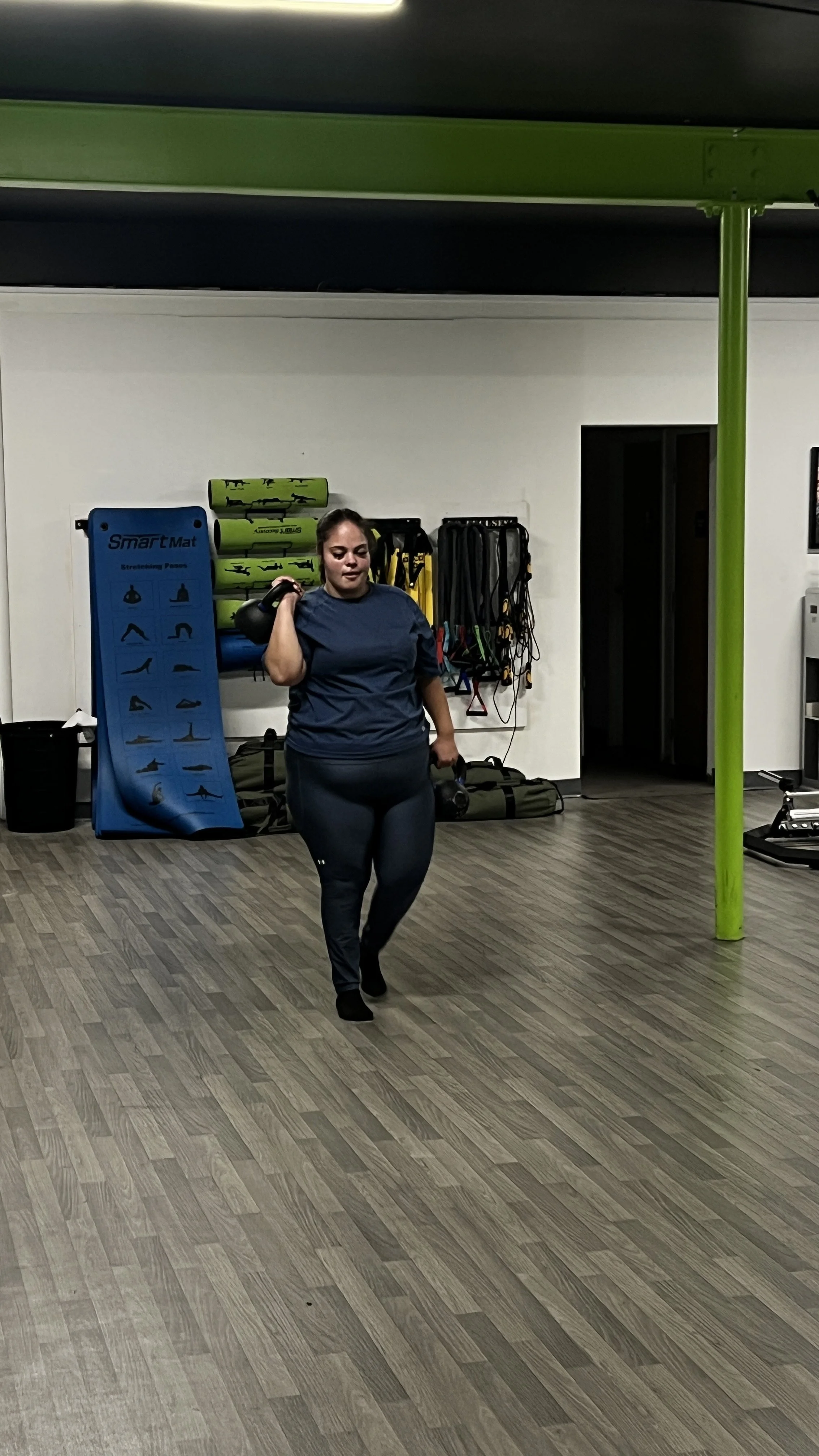 A woman in workout clothes walking inside a gym, carrying a kettlebell on her shoulder and holding a piece of equipment in her other hand.