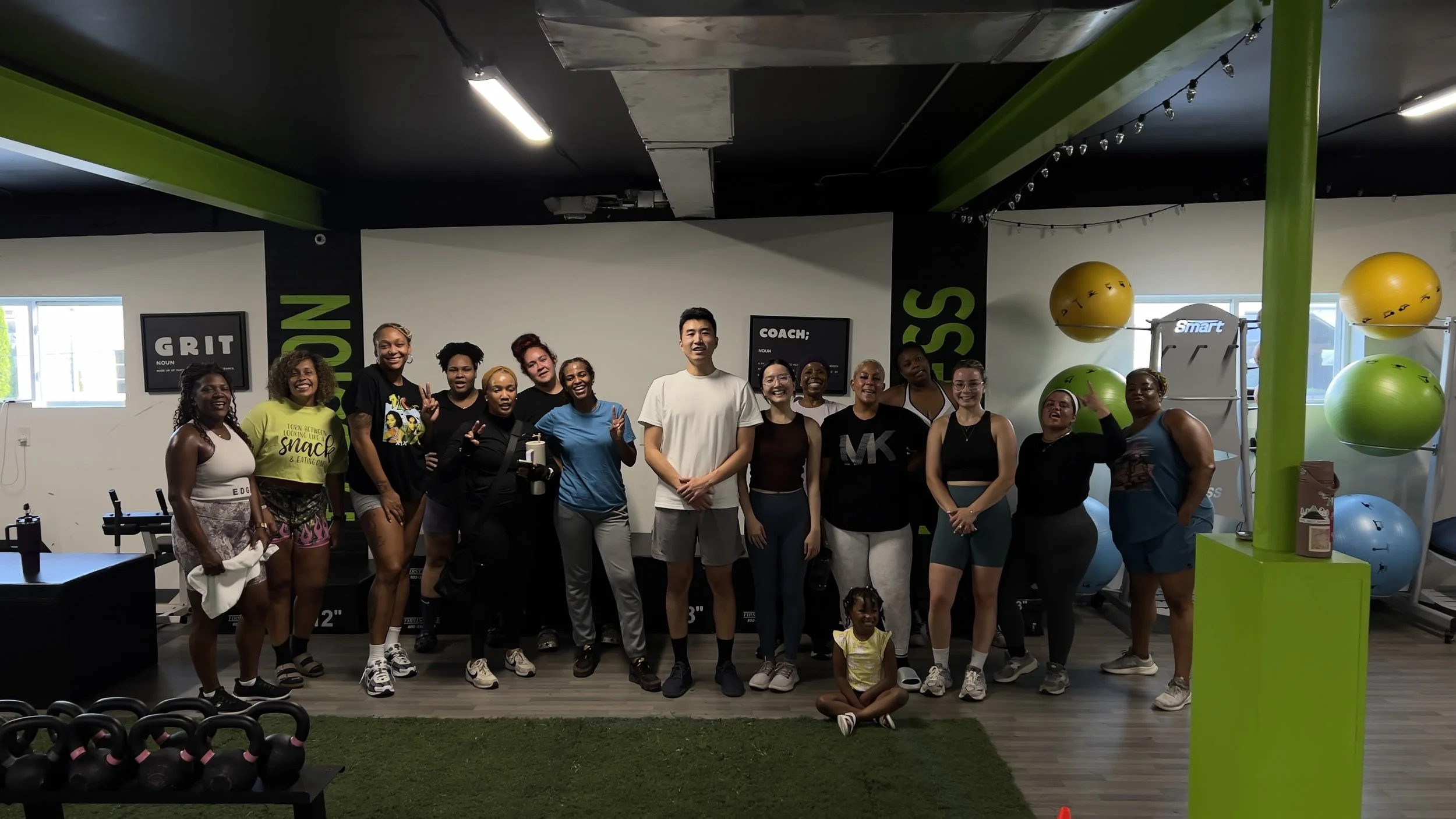 A group of diverse women and children posing together in a gym with green fitness equipment and motivational signs, including one that says 'GRIT'.
