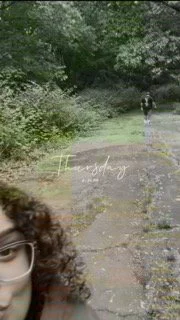 Part of a woman's face with glasses in the bottom left corner, blurred background of a green park path with trees and a person walking in the distance.