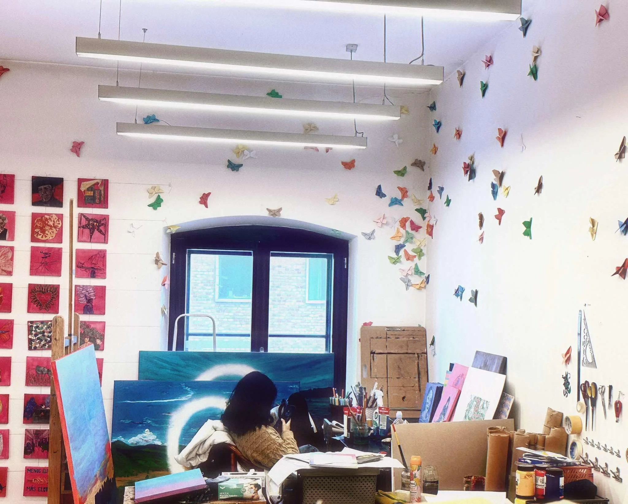 An art studio with colorful origami butterflies decorating the wall, several paintings, art supplies, and a person working on an artwork.