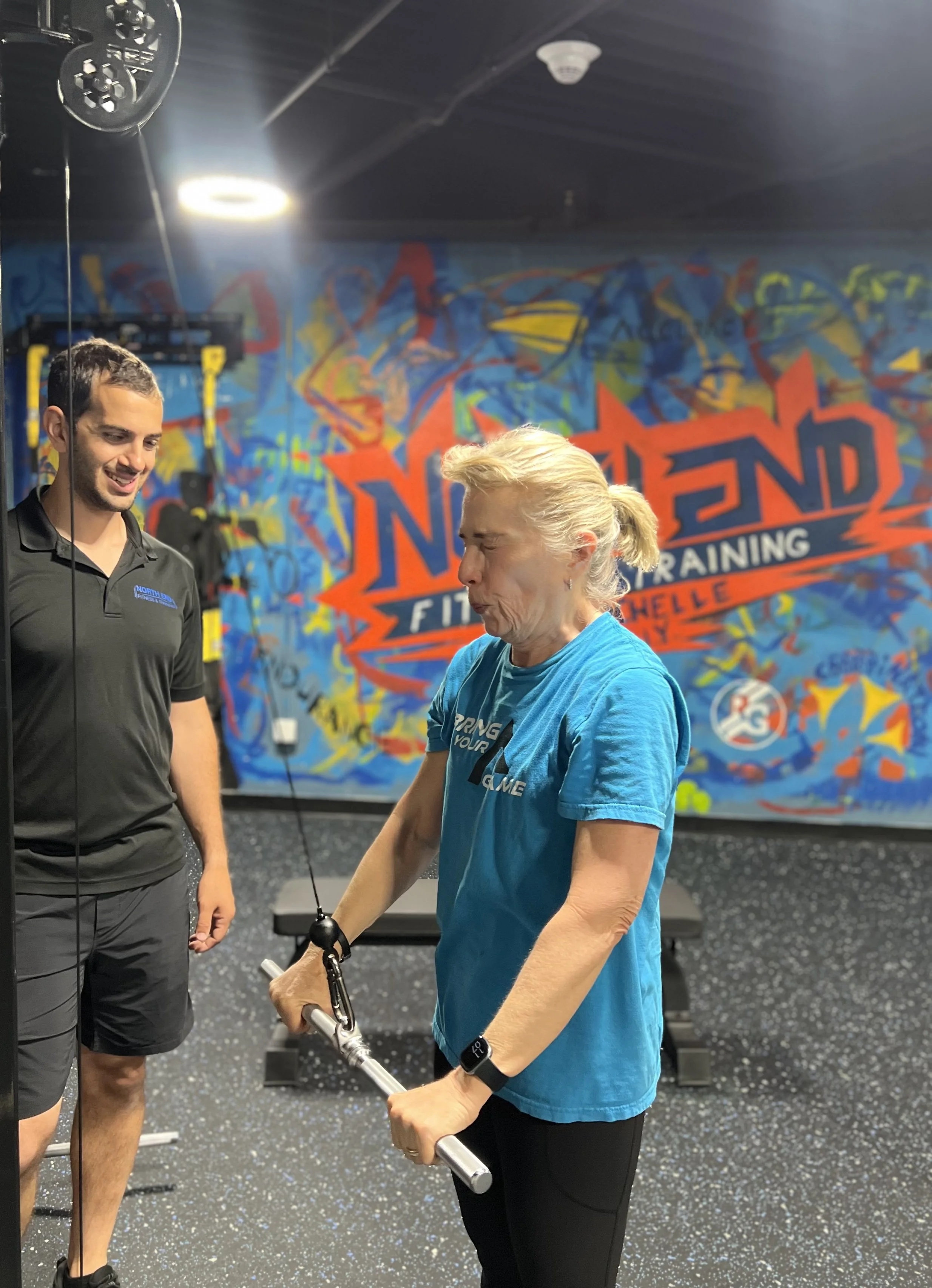 A senior woman in a blue shirt holding a barbell with a trainer at a gym with colorful graffiti wall in the background.