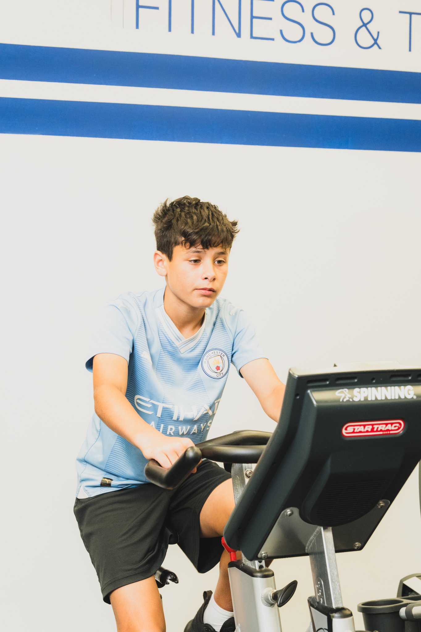 A young man in a light blue Manchester City soccer jersey and black shorts is riding a stationary exercise bike in a gym. He appears to be focused on the screen in front of him, which is part of the exercise equipment.