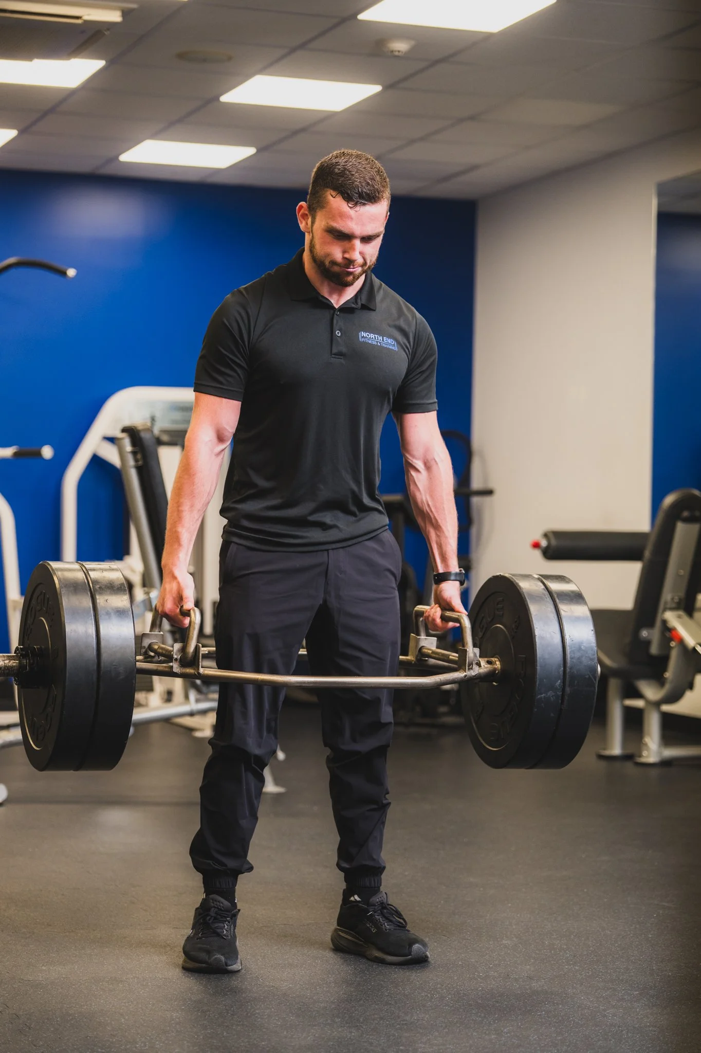 A man working out with dumbbells in a gym, wearing a black polo shirt with 'North End' logo, black pants, and black shoes.