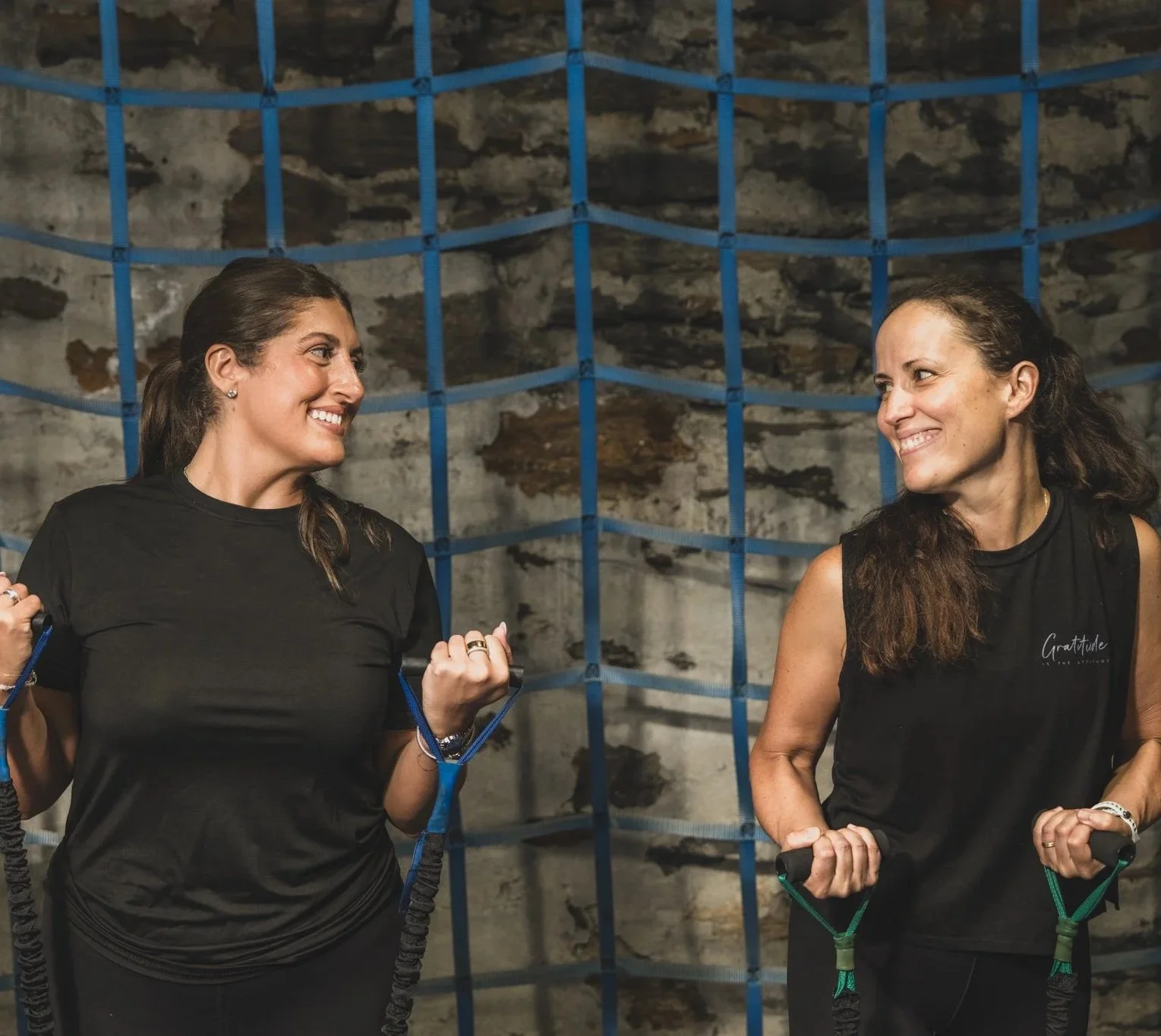 Two women smiling and looking at each other, holding resistance bands in a gym with a rocky wall and blue grid in the background.