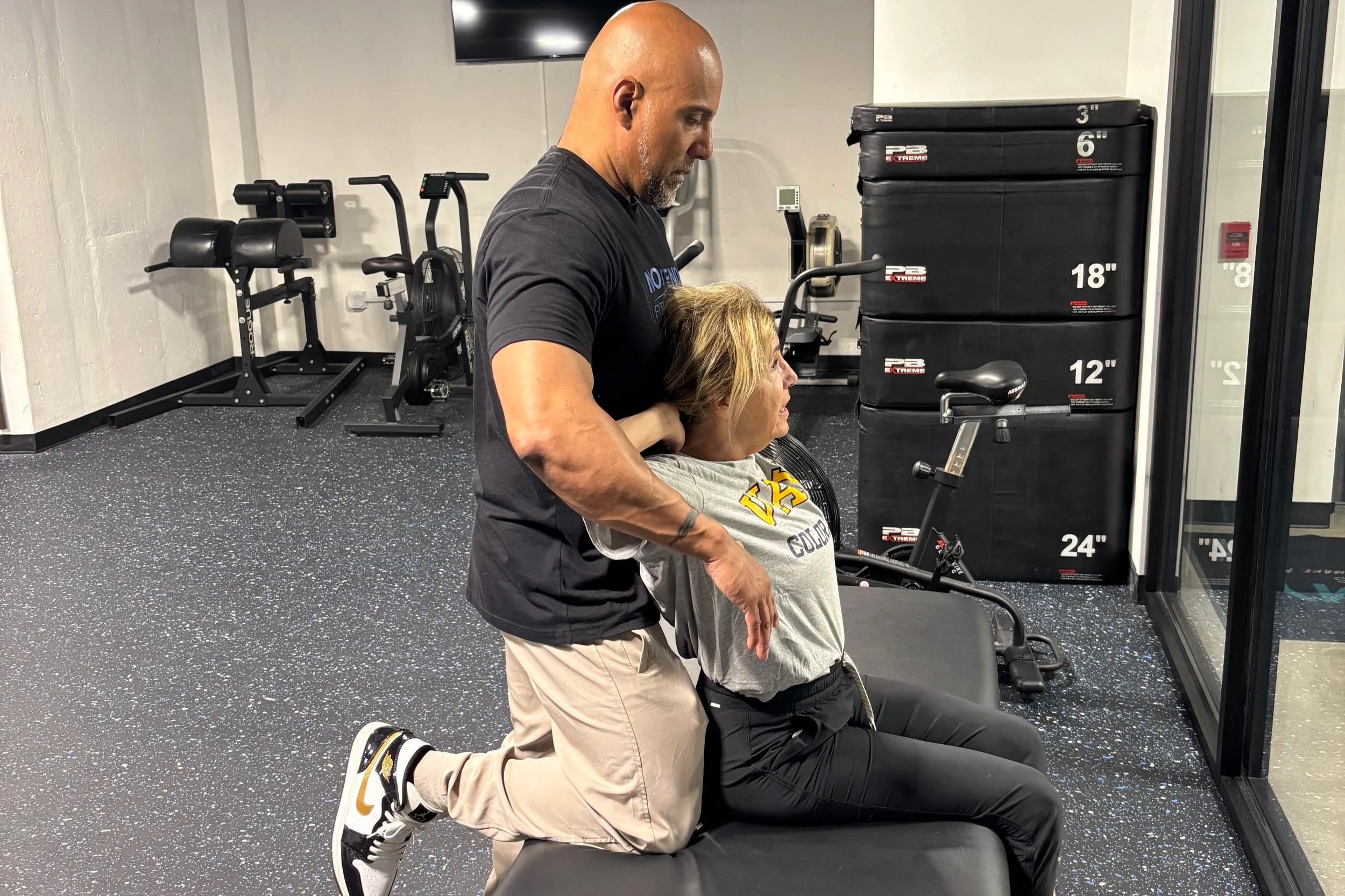 A man is kneeling and assisting a woman in a wheelchair during a workout session in a gym. The woman appears to be performing an exercise, with her arms on the wheelchair's armrests. The gym has exercise equipment such as a rowing machine and stacked workout pads in the background.