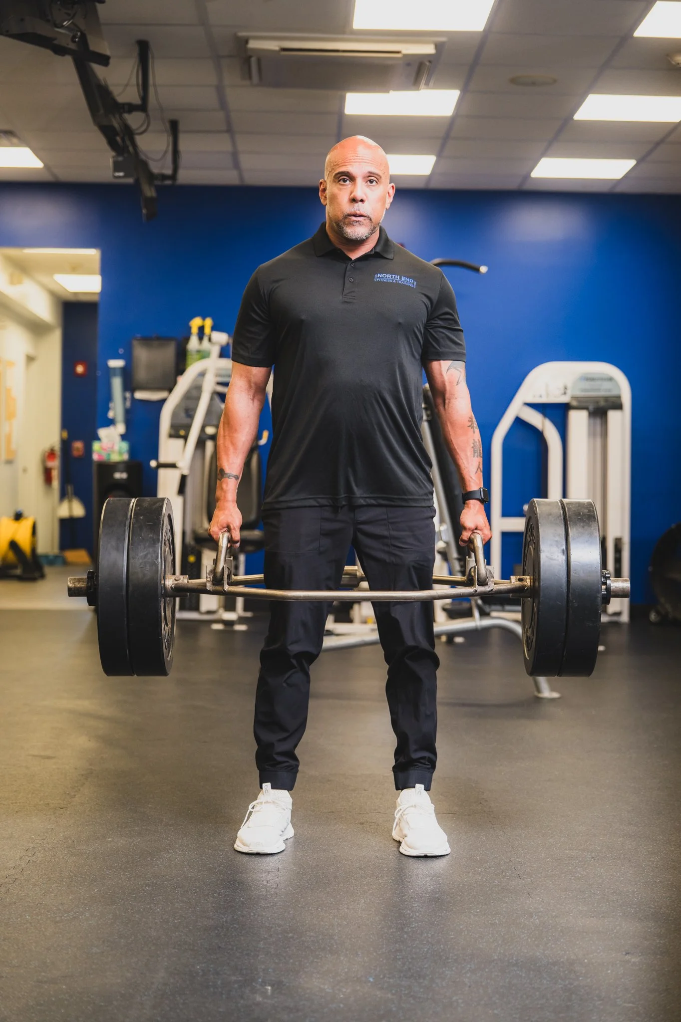 A man with a bald head and tattoos on his arms is standing in a gym, holding a barbell with large weights.