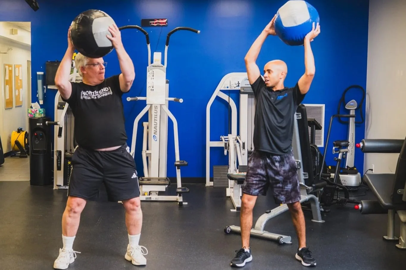 Two men lifting medicine balls in a gym with exercise equipment and blue walls.