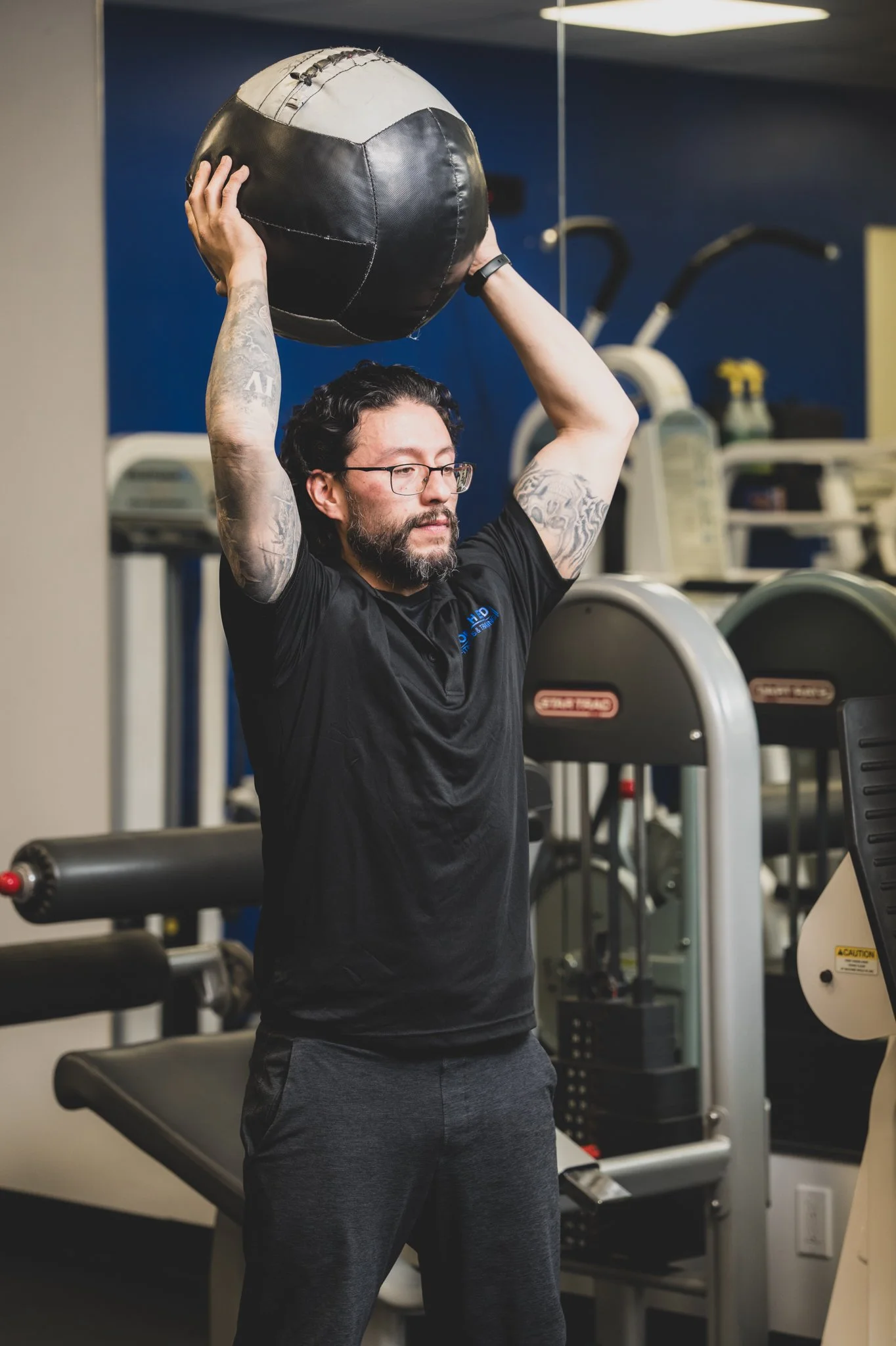 A man with tattoos and glasses in a gym holds a large black and white medicine ball above his head. The gym has various exercise machines and equipment in the background.