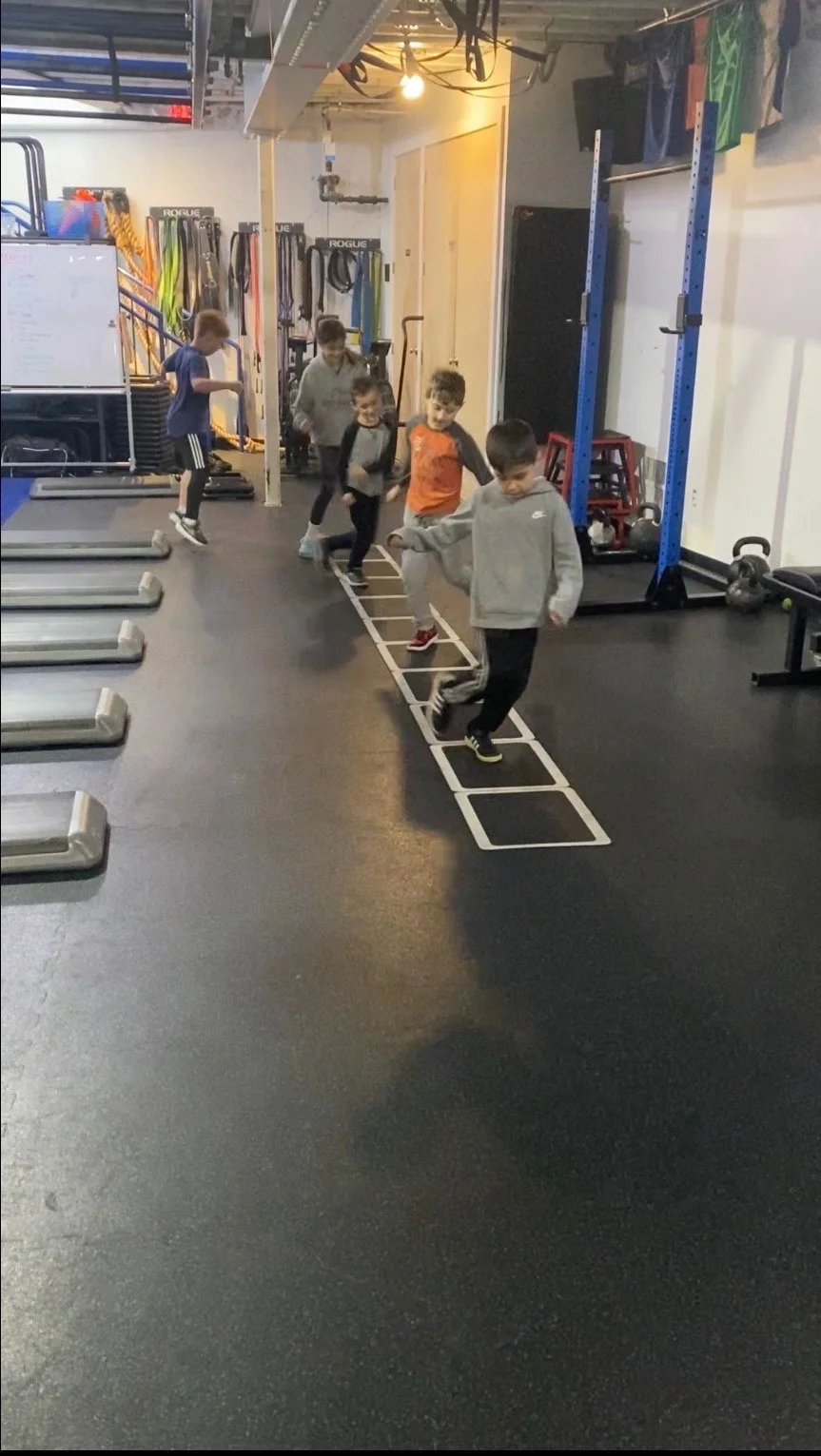 A group of kids playing hopscotch on a black gym floor inside a fitness or gym facility, with exercise equipment and colorful resistance bands visible in the background.