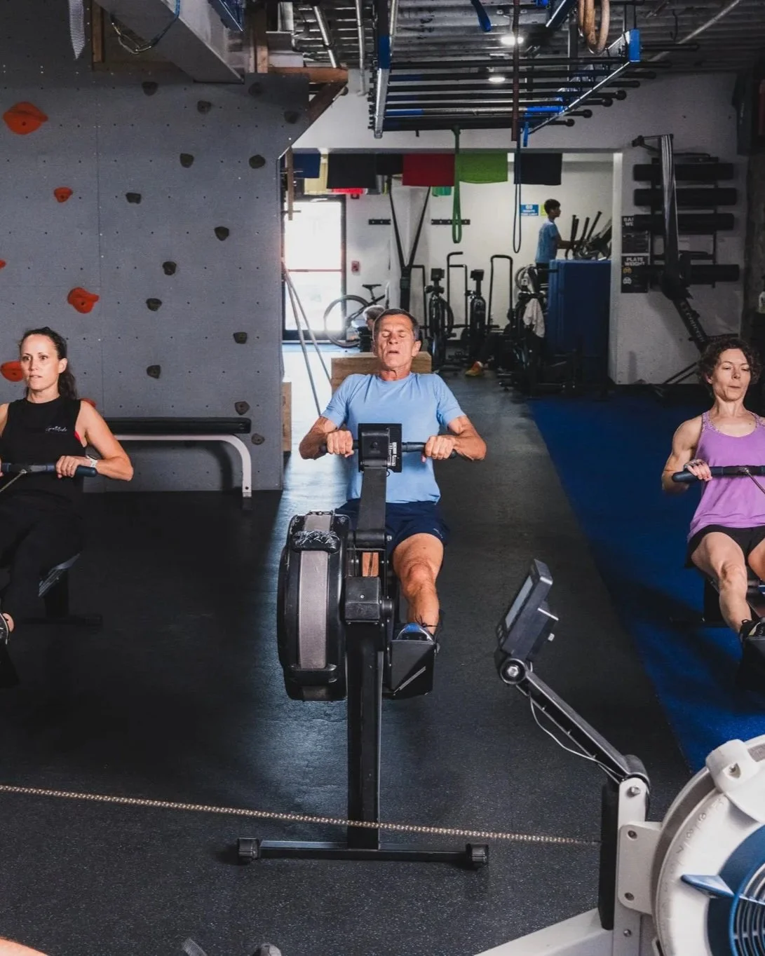 People working out on rowing machines in a gym, with a rock climbing wall and exercise equipment in the background.