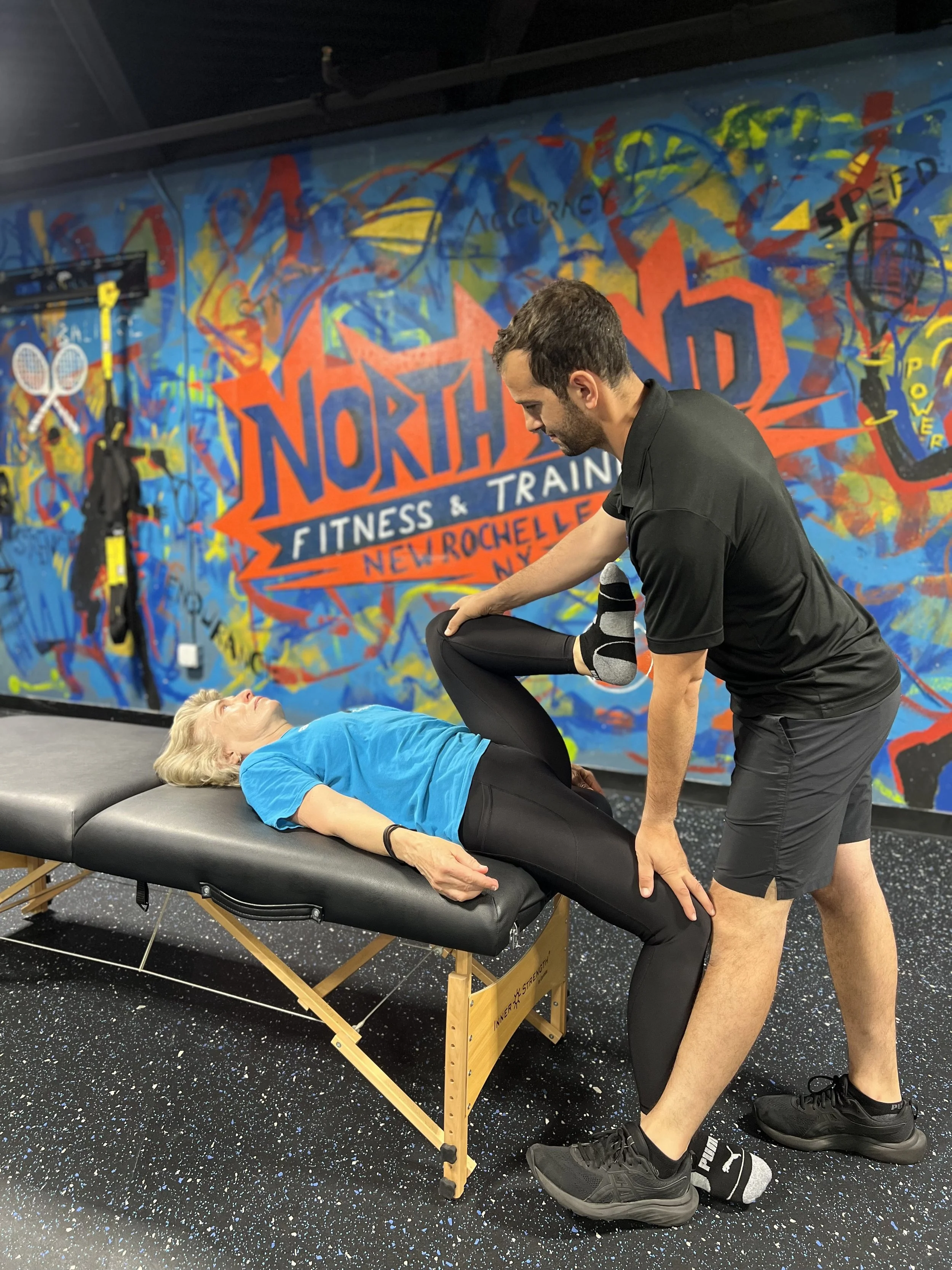 A male fitness trainer assisting an older woman with a stretching exercise on a massage table at a fitness center with colorful wall art and gym equipment in the background.
