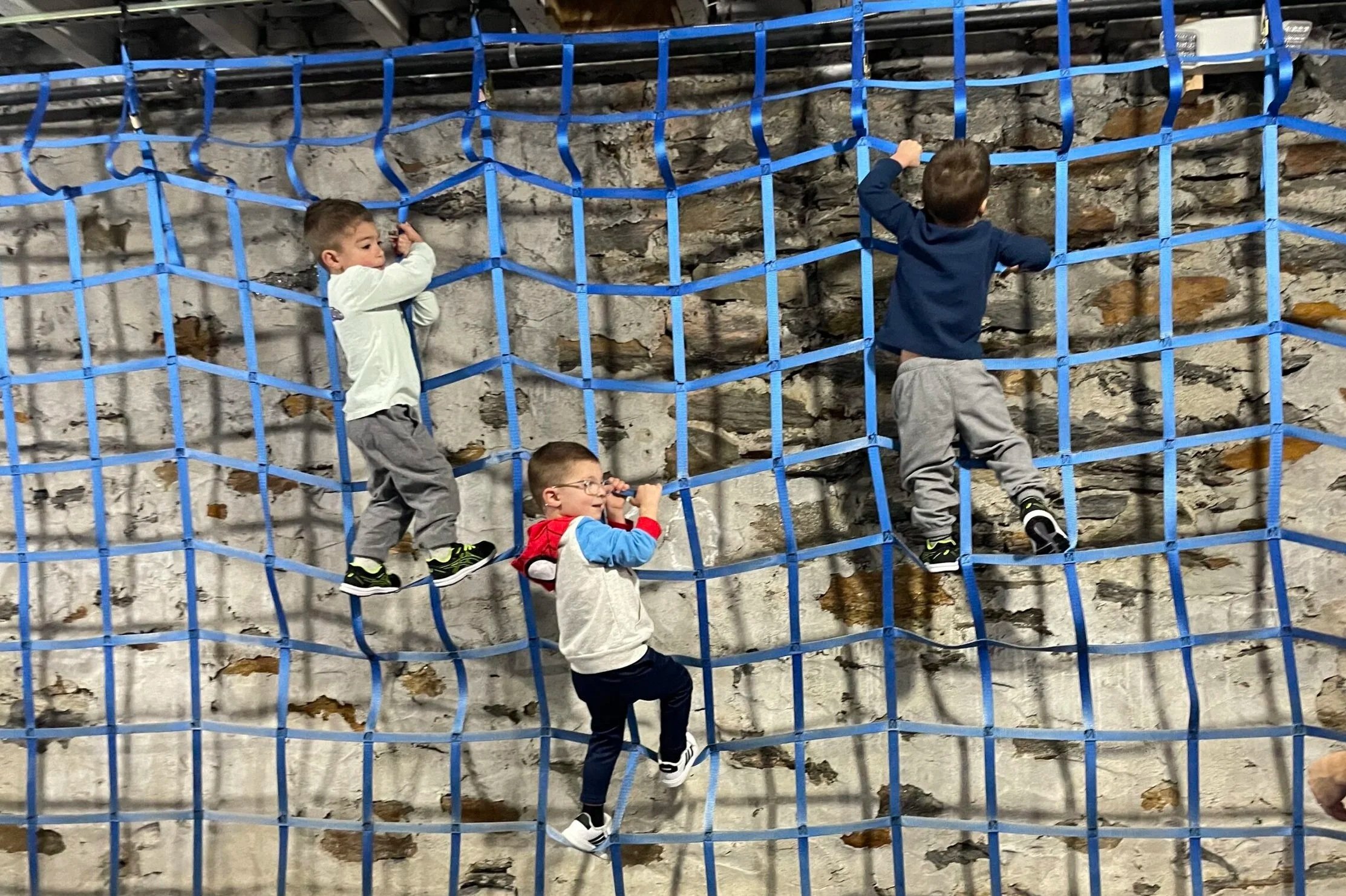 Four young boys climbing on a blue rope net attached to a stone wall indoor.