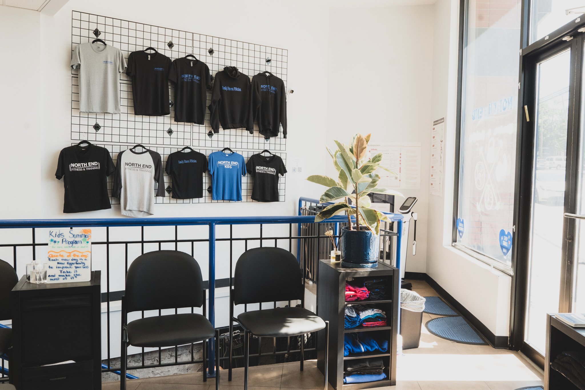 Display of North End branded t-shirts and hoodies on a grid wall inside a bright store, with a table and chairs in front and a potted plant near a glass door.