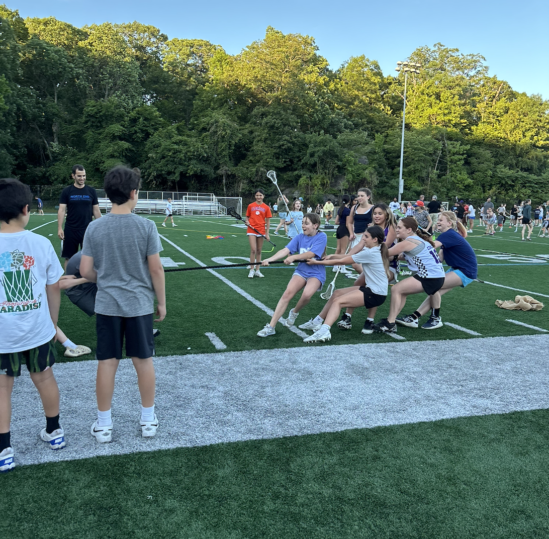 Group of children playing tug of war on a sports field with adults and other children observing in the background, surrounded by trees under a clear blue sky.