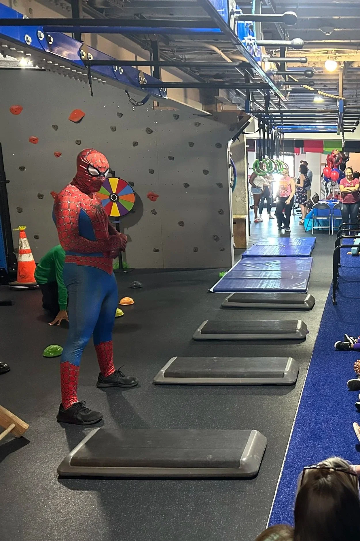 A room with a Spider-Man costume standing in front of a rock climbing wall with orange holds. There are several black aerobic step platforms on the floor, and a group of people in the background, some standing near a table with balloons and others engaging in activities.