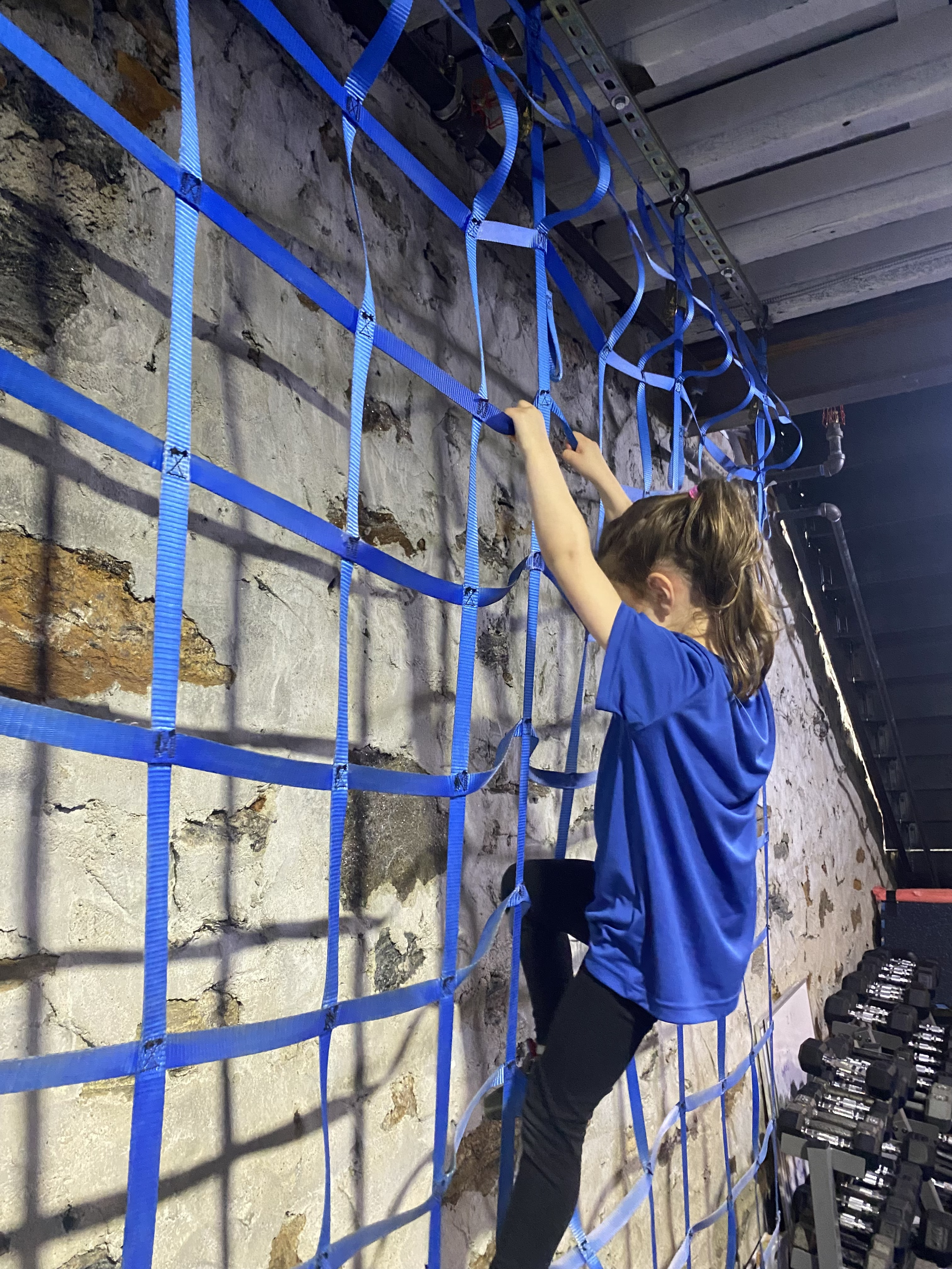 A woman in a blue shirt and black pants is doing an exercise behind a blue safety net in a gym. Dumbbells are visible on a rack adjacent to the net.