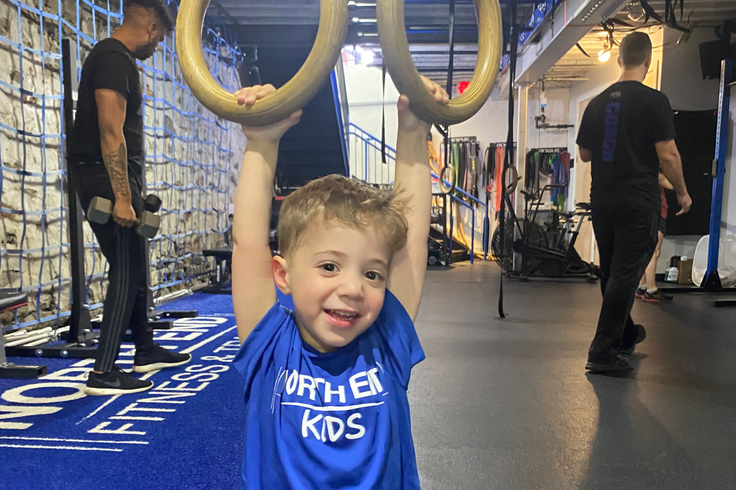 A young boy smiling, holding onto gymnastic rings above his head in a gym. Two trainers or staff members are visible in the background, one on the left holding a dumbbell and the other on the right, both in black shirts. The gym has workout equipment, rings, and a blue mat with white writing.
