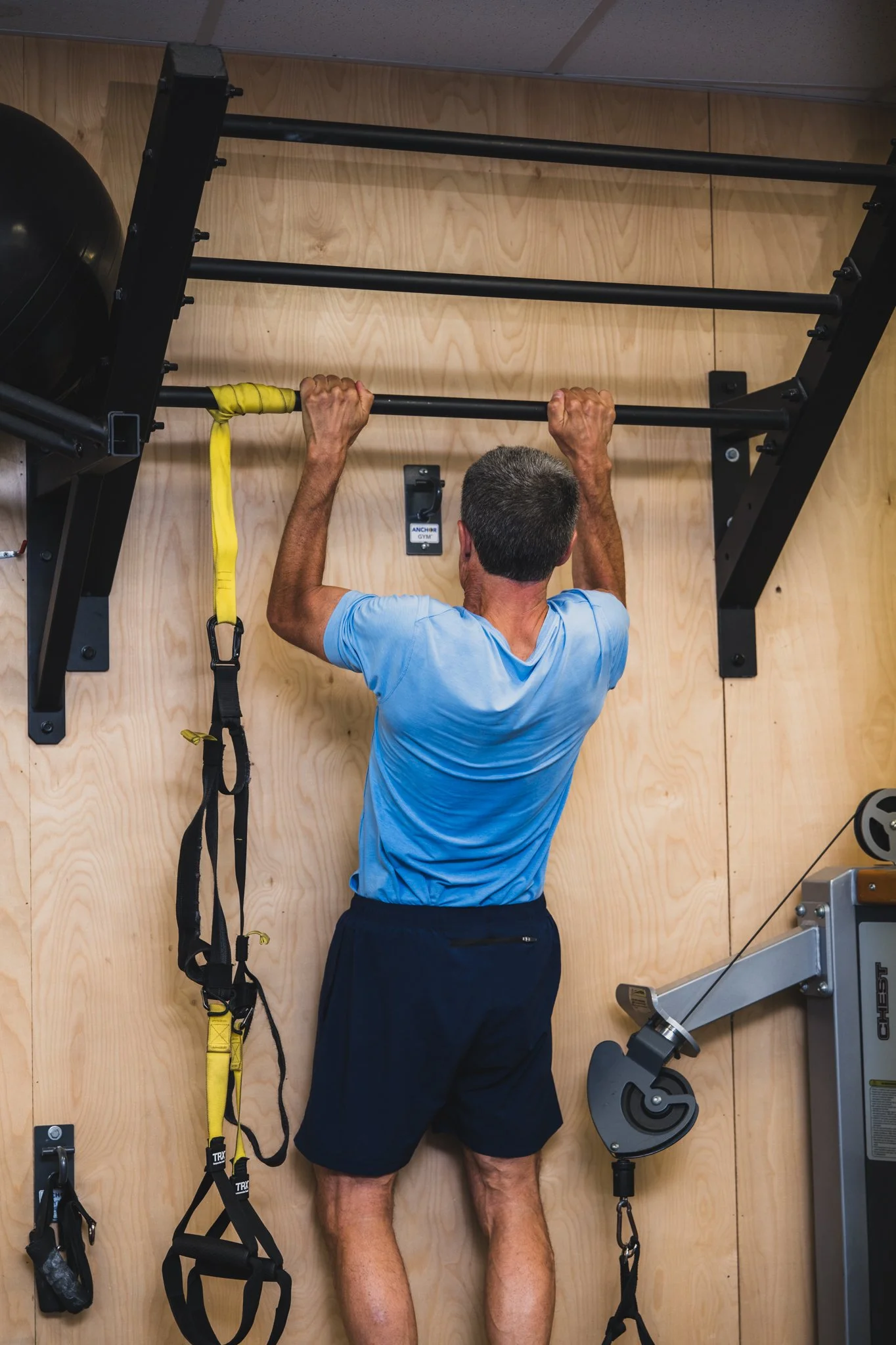 A man in a blue shirt and black shorts doing pull-ups on a black pull-up bar in a gym with a wood-paneled wall.