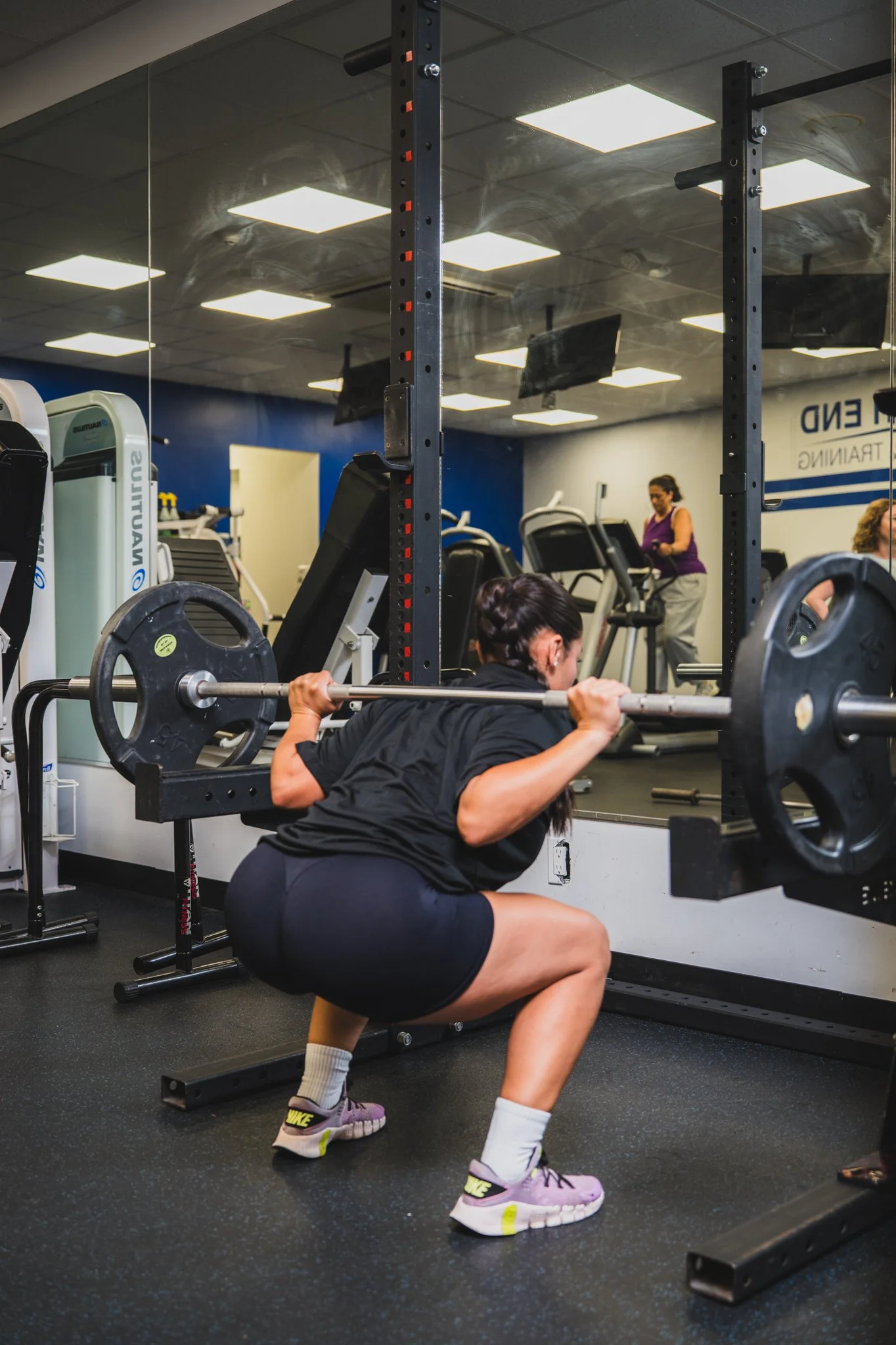 A woman in black workout clothes performing a squat with a barbell in a gym, with other people working out in the background.