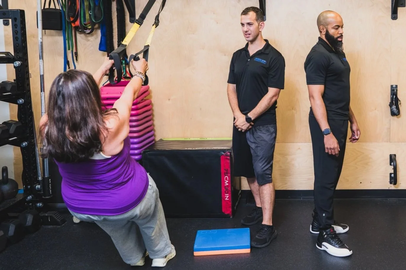 Woman performing suspension training exercise with staff observing in a gym.