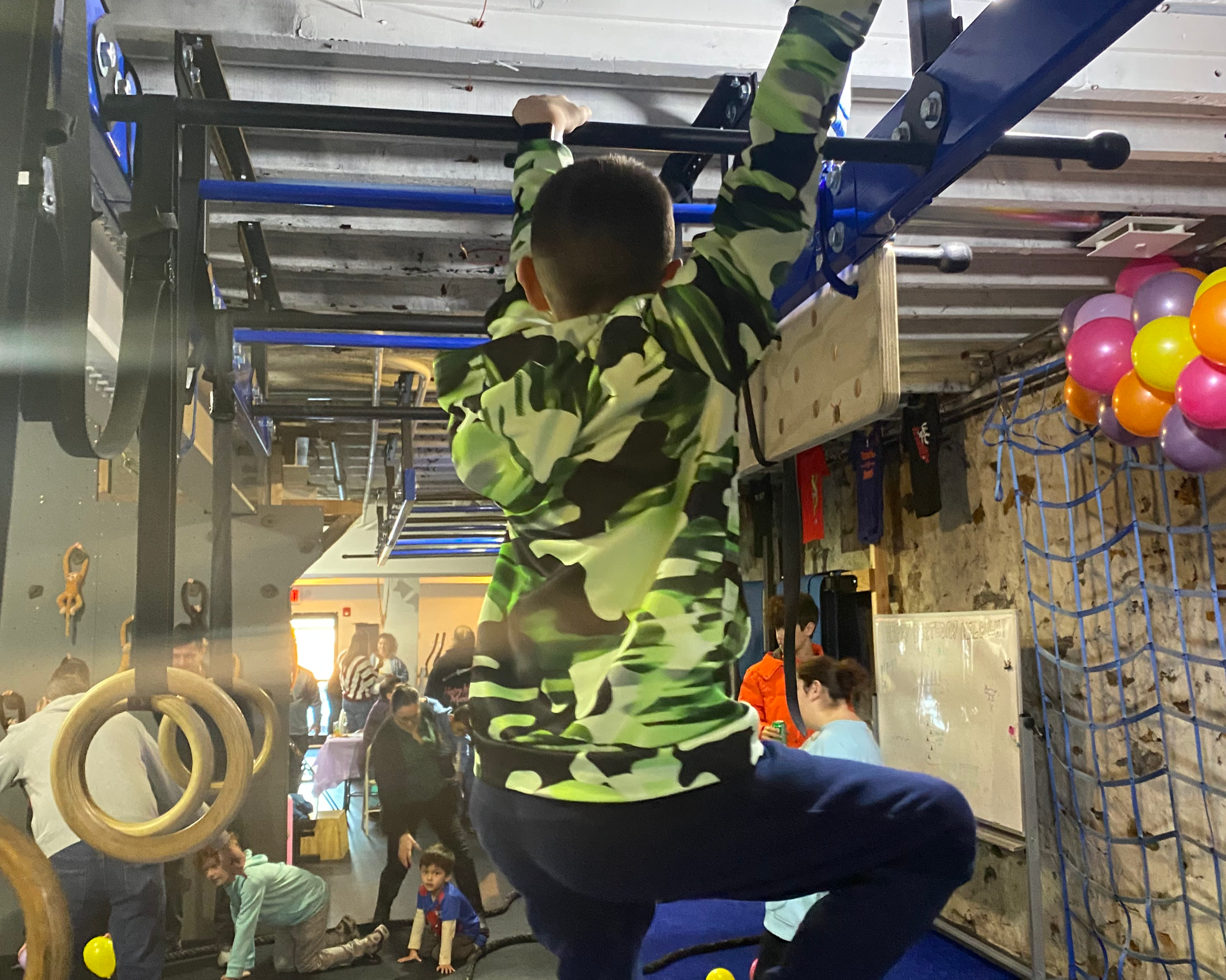 Child in camouflage hoodie climbing on gym equipment in indoor play area with colorful balloons and other children and adults in background.