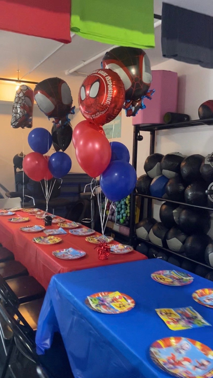 Party decorations with red, blue, and Spider-Man themed balloons, party plates, and colorful tablecloths, in a gym or activity room with workout equipment on shelves.