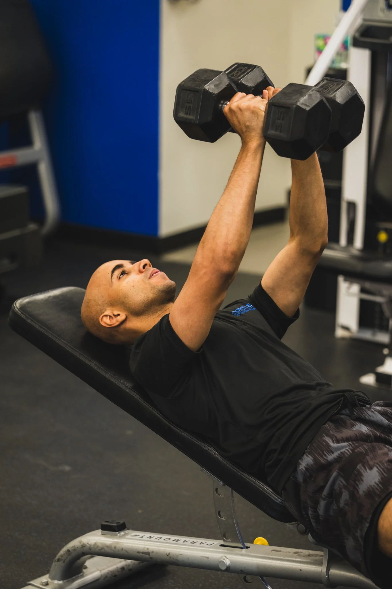 A man is lying on a workout bench, lifting two dumbbells in a gym. He has a bald head, and is wearing a black t-shirt and athletic shorts. The background includes gym equipment and a blue wall.