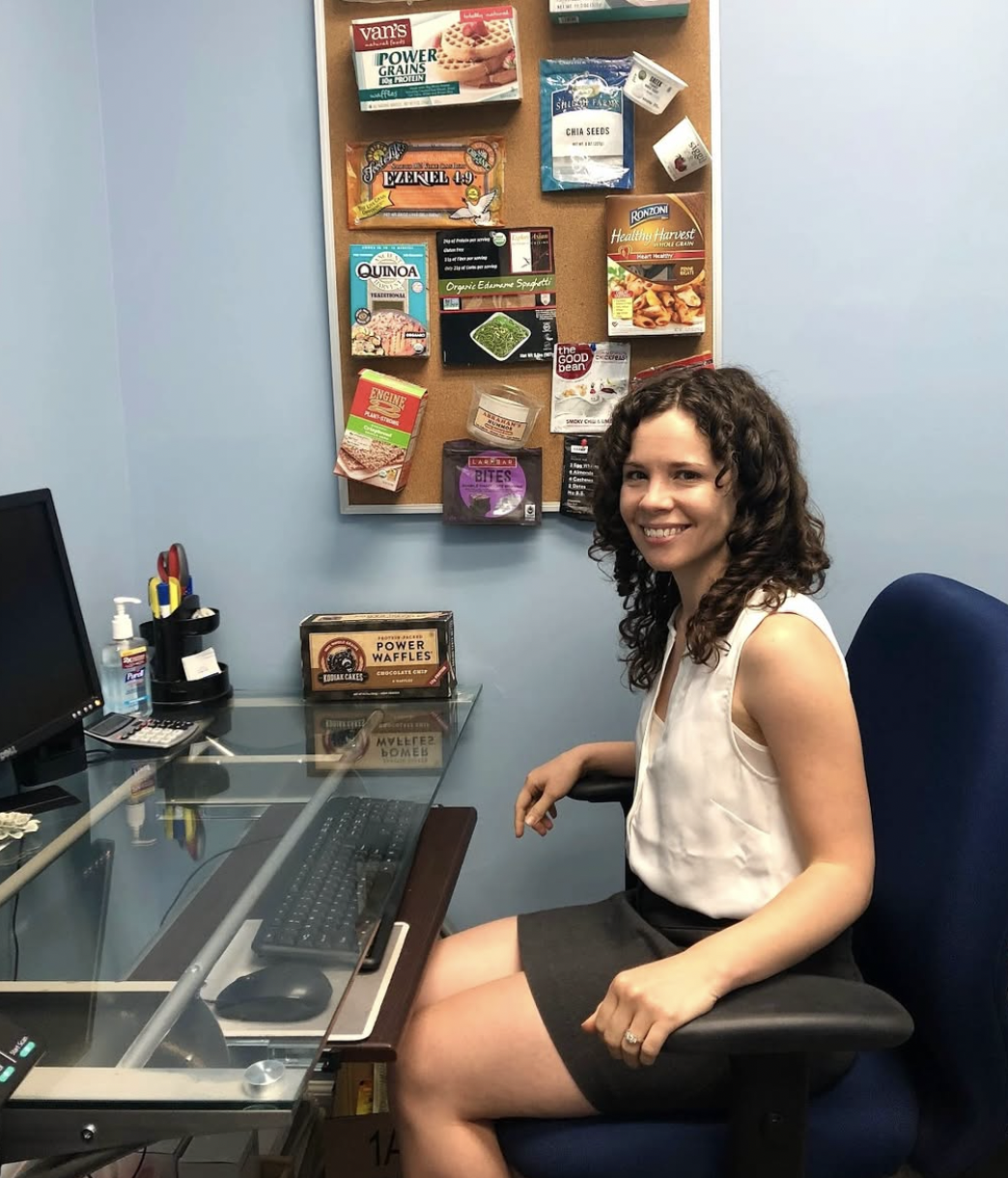 A woman seated in an office chair at a glass desk with a computer monitor, keyboard, and mouse, smiling at the camera. Behind her is a bulletin board with various snack packets and food items pinned to it.