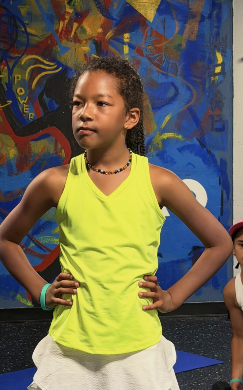 A young girl with curly hair standing with her hands on her hips in front of a colorful mural. She is wearing a bright yellow tank top, a beaded necklace, and a teal bracelet.