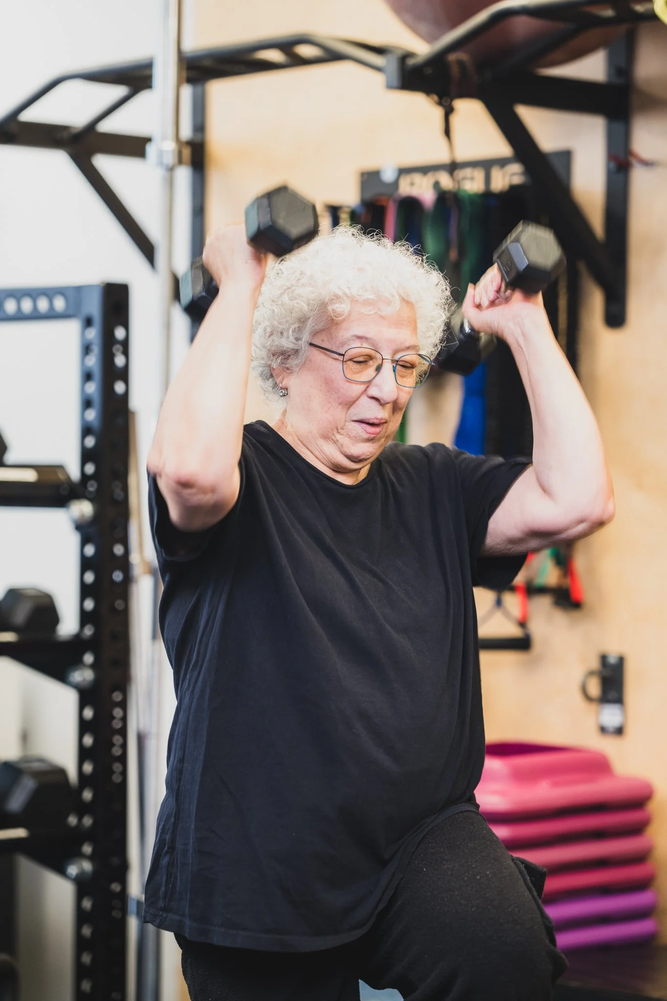 A senior with curly white hair and glasses is lifting black dumbbells in a gym. She appears focused on her workout, standing in front of a wall with various exercise equipment and fitness accessories visible in the background.