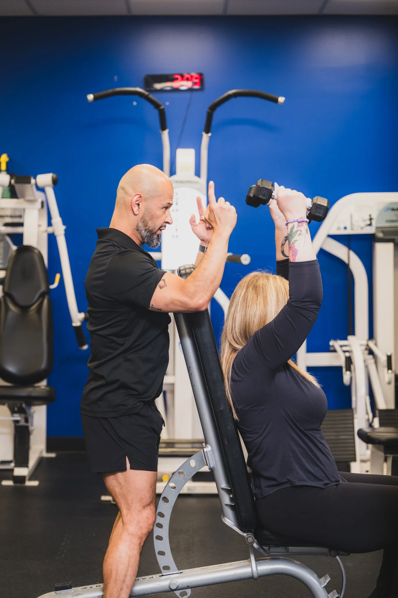 A man with a bald head and gray beard helping a woman with blonde hair seated in a gym. The man is giving instructions or encouragement while the woman is doing a seated exercise. The gym has blue walls, fitness machines, and a digital clock in the background.
