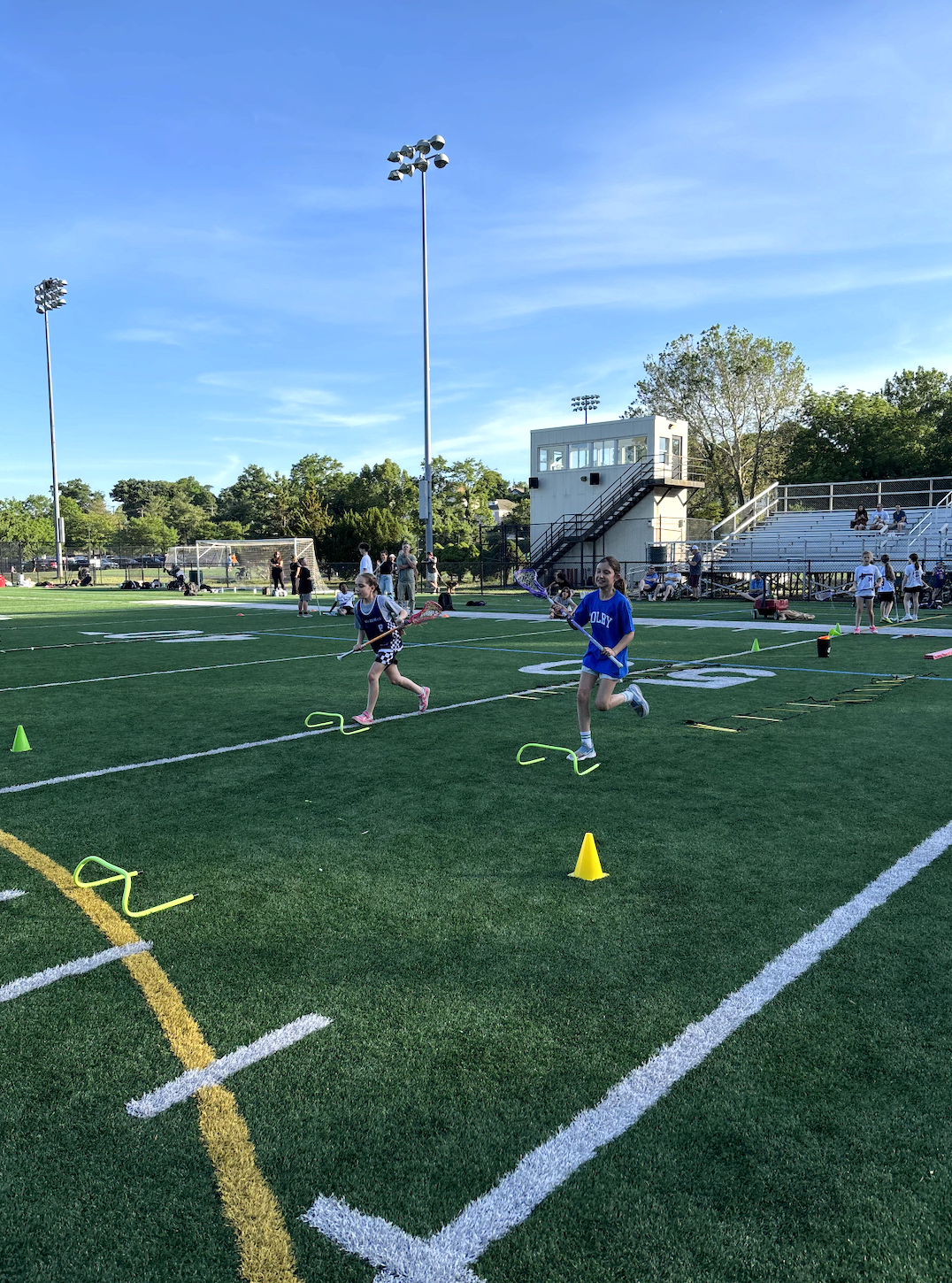 Children participating in a field hockey drill on a sports field, with cones and hurdles set up under a clear blue sky, and spectators in the bleachers.