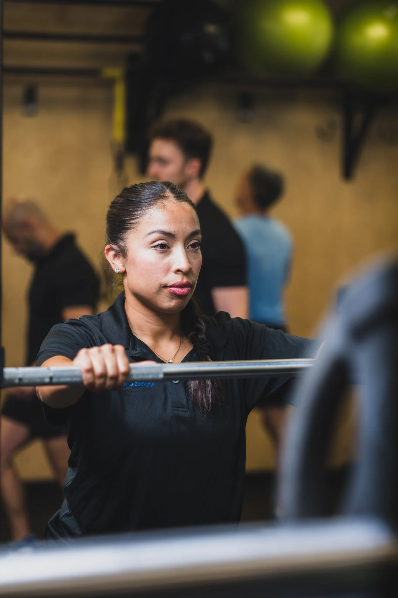 A woman with dark hair tied back, wearing a black shirt, appears to be lifting a barbell during a workout at the gym. In the background, blurred figures of other people are visible, along with gym equipment and green balls on a rack.