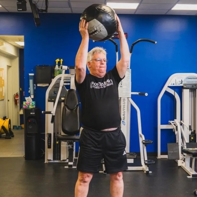 Older man with glasses lifting a black and gray medicine ball overhead in a gym with blue walls and workout equipment.